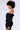 A woman with curly hair is wearing the IN YOUR DREAMS RUFFLE MINI DRESS, a black, off-the-shoulder LBD featuring ruffle detailing and sheer long sleeves. She is standing in profile against a plain background, accessorizing her stylish look with large, rectangular earrings.