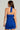 A woman with long brown hair is wearing the SERPENTI PLUNGE MINI DRESS IN BLUE, a halter-neck, backless design that exudes elegance. In her left hand, she holds a small textured clutch. The sleek bodycon party dress fits her perfectly and is accentuated by a discreet zip at the back against a plain gray background.