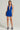 A person stands against a plain background, dressed in the Serpenti Plunge Mini Dress in Blue, featuring a deep V-neckline. They hold a clutch in one hand and wear platform heels, exuding effortless elegance with their neutral expression.