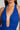 A close-up view of an individual wearing the vibrant SERPENTI PLUNGE MINI DRESS IN BLUE, featuring a deep V-neckline. The fabric is ruched with small, subtle sparkles. The person has long brown hair and is accessorized with large, elegant earrings.