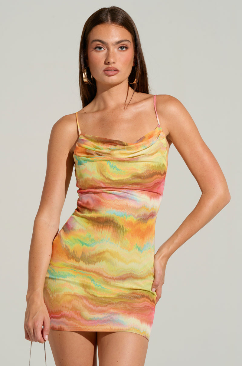 A woman confidently models the CLARA PRINTED MINI DRESS, showcasing its striking technicolor swirl pattern. This sleeveless bodycon design is highlighted by thin straps and a cowl neckline. She accessorizes with hoop earrings, her long straight hair contrasting against a neutral gray background.