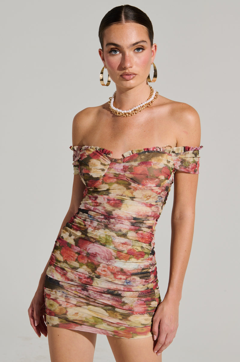 A woman poses in the ELODI floral print ruched mini dress with an off-shoulder bodycon silhouette. She accessorizes with large hoop earrings and a layered necklace, her hair is sleekly styled back as she stands against a plain background, gazing directly at the camera.