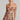 A woman poses in the ELODI floral print ruched mini dress with an off-shoulder bodycon silhouette. She accessorizes with large hoop earrings and a layered necklace, her hair is sleekly styled back as she stands against a plain background, gazing directly at the camera.