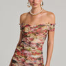A woman poses in the ELODI floral print ruched mini dress with an off-shoulder bodycon silhouette. She accessorizes with large hoop earrings and a layered necklace, her hair is sleekly styled back as she stands against a plain background, gazing directly at the camera.