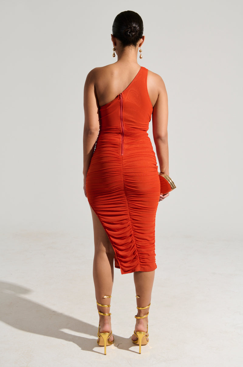 A woman in a fitted Nora Cutout Ruched Midi Dress in orange is standing with her back to the camera. She holds a small clutch in her left hand and is wearing strappy yellow high heels. Her hair is tied back in a sleek bun, accentuating the one-shoulder design against the plain, light grey background.