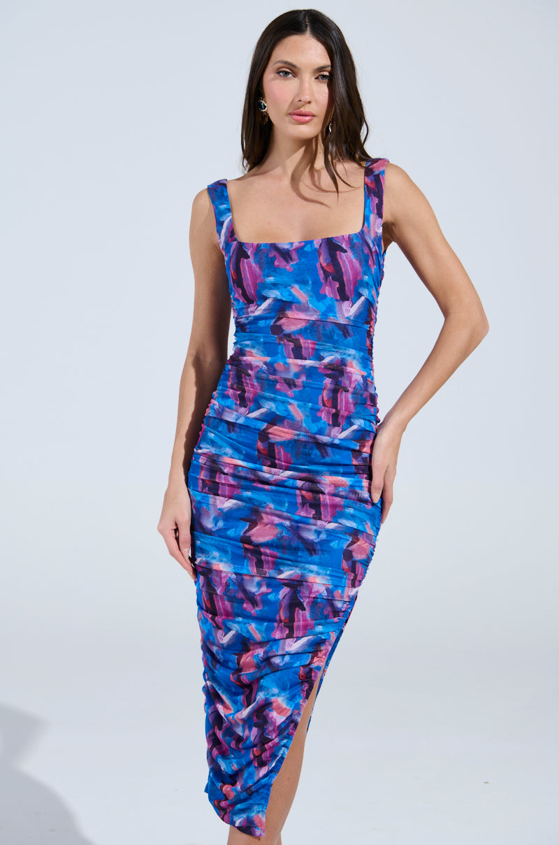 A woman radiates confidence in the YOU MAKE ME BLUSH ABSTRACT MIDI DRESS, featuring a vivid blue and purple pattern. This form-fitting midi dress, made from stretchy mesh with a square neckline and side slit, is elegantly showcased against a simple, light-colored backdrop.