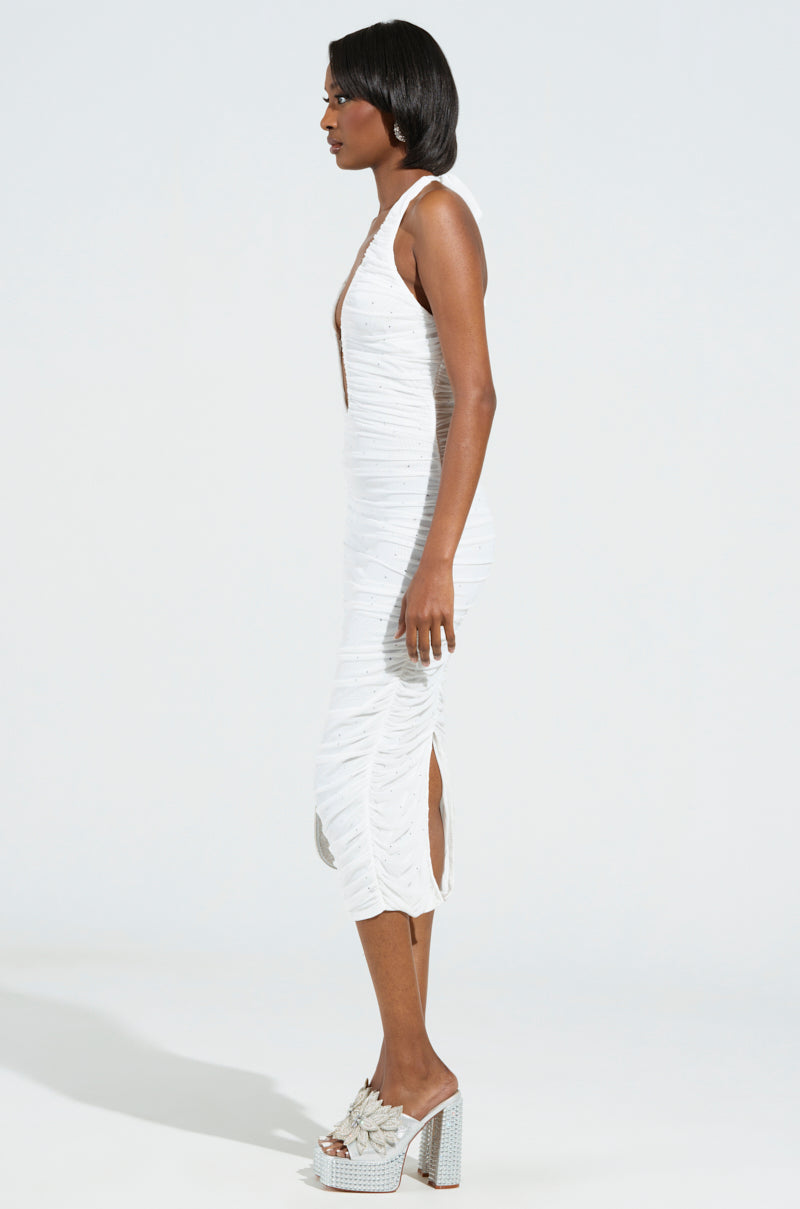 An individual wearing the sleeveless Serpenti Plunge Midi Dress in Ivory, showcasing a deep neckline and side slit, stands in profile against a plain backdrop. The dress is adorned with rhinestone embellishments and is paired with high-heeled sandals that feature fringe detailing.