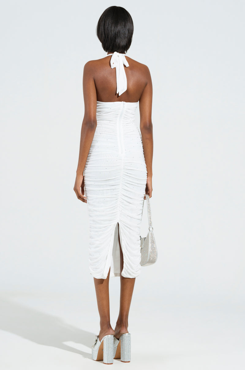 A person dazzles in the Serpenti Plunge Midi Dress in Ivory, showcasing a halter-neck design adorned with rhinestone embellishments and a ruched construction. They hold a sparkling silver handbag and wear platform heels while standing against a plain, light-colored background.