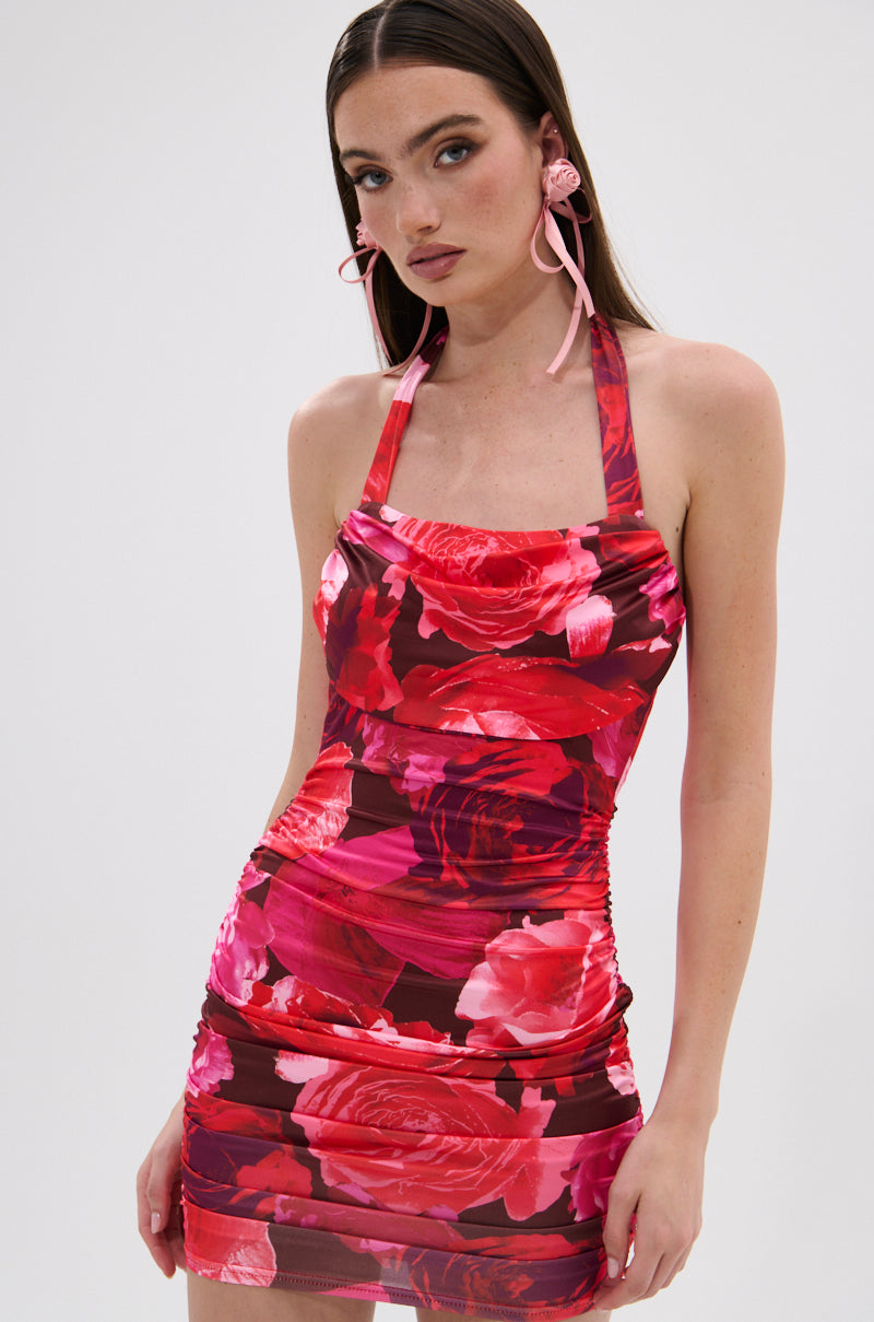 A woman with long brown hair wears the POSIE PRINTED MINI DRESS, a curve-hugging style with bold red and pink florals. She accessorizes with large pink flower earrings and poses against a plain white background.