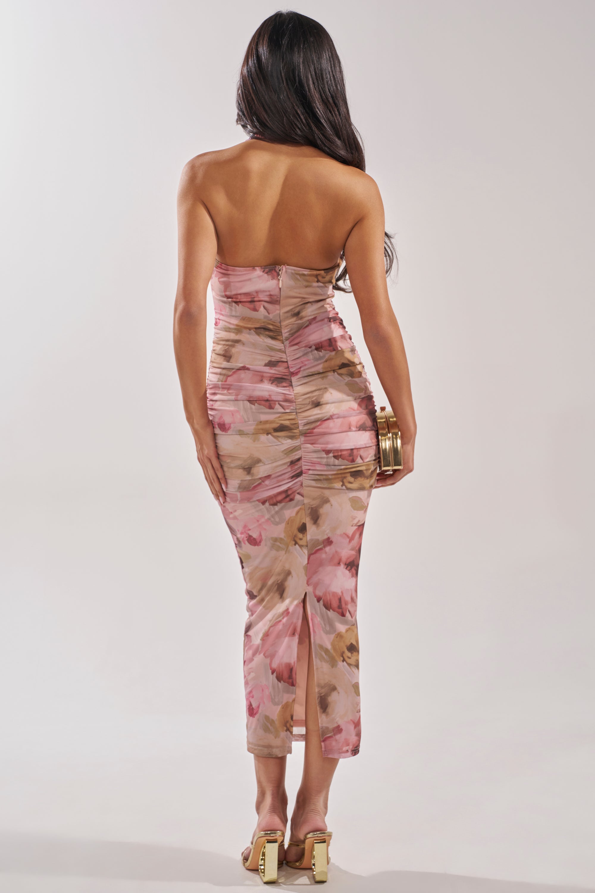 A woman with long dark hair, seen from behind, wears the I DON'T CHASE FLORAL MAXI DRESS in pink and beige with high-heeled sandals and a gold clutch—an ideal chic garden party look. 3