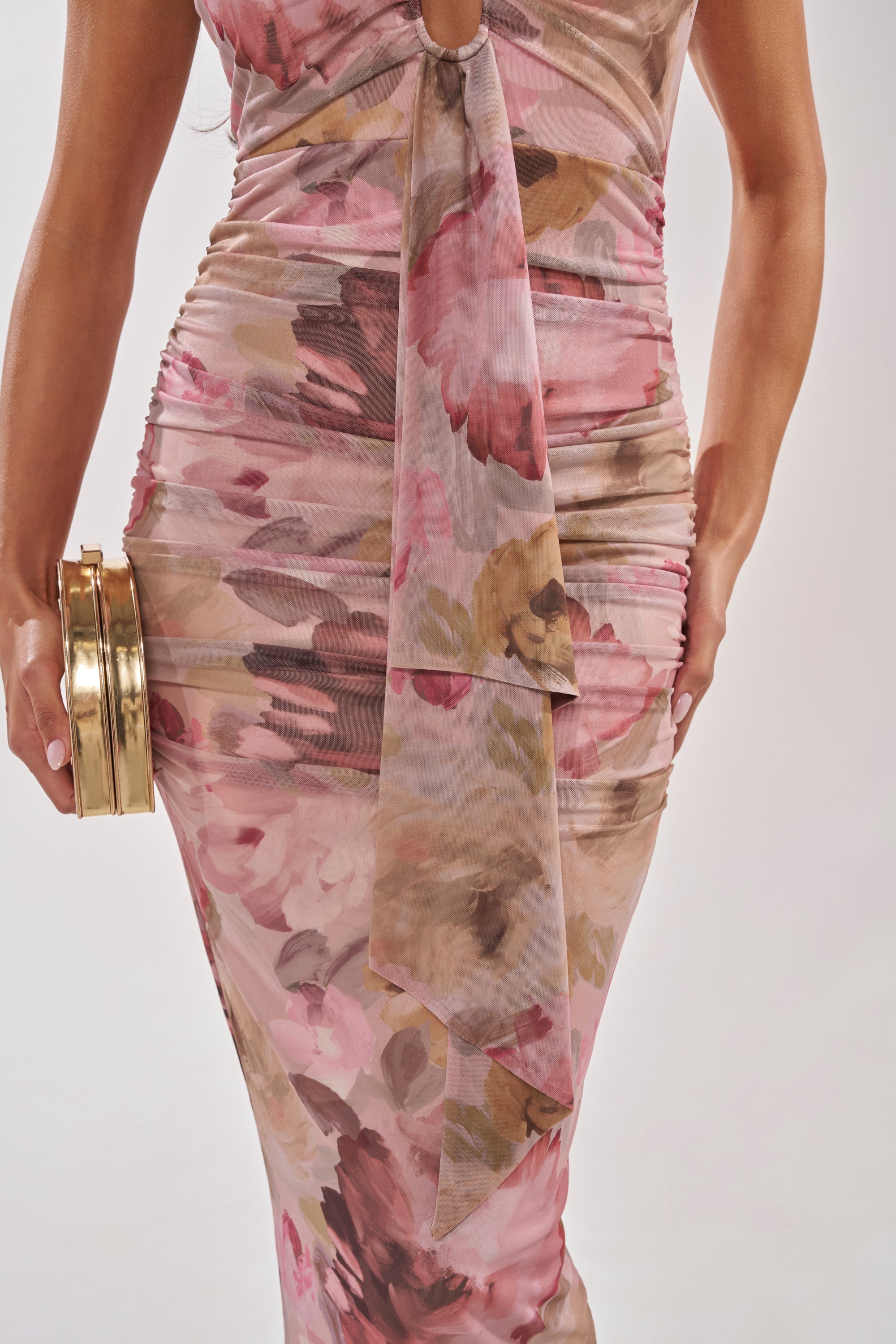 Wearing the I DON'T CHASE FLORAL MAXI DRESS, a fitted sleeveless garden party look with pink and brown florals, a knotted center detail, and front draping, paired with a round gold clutch. 5