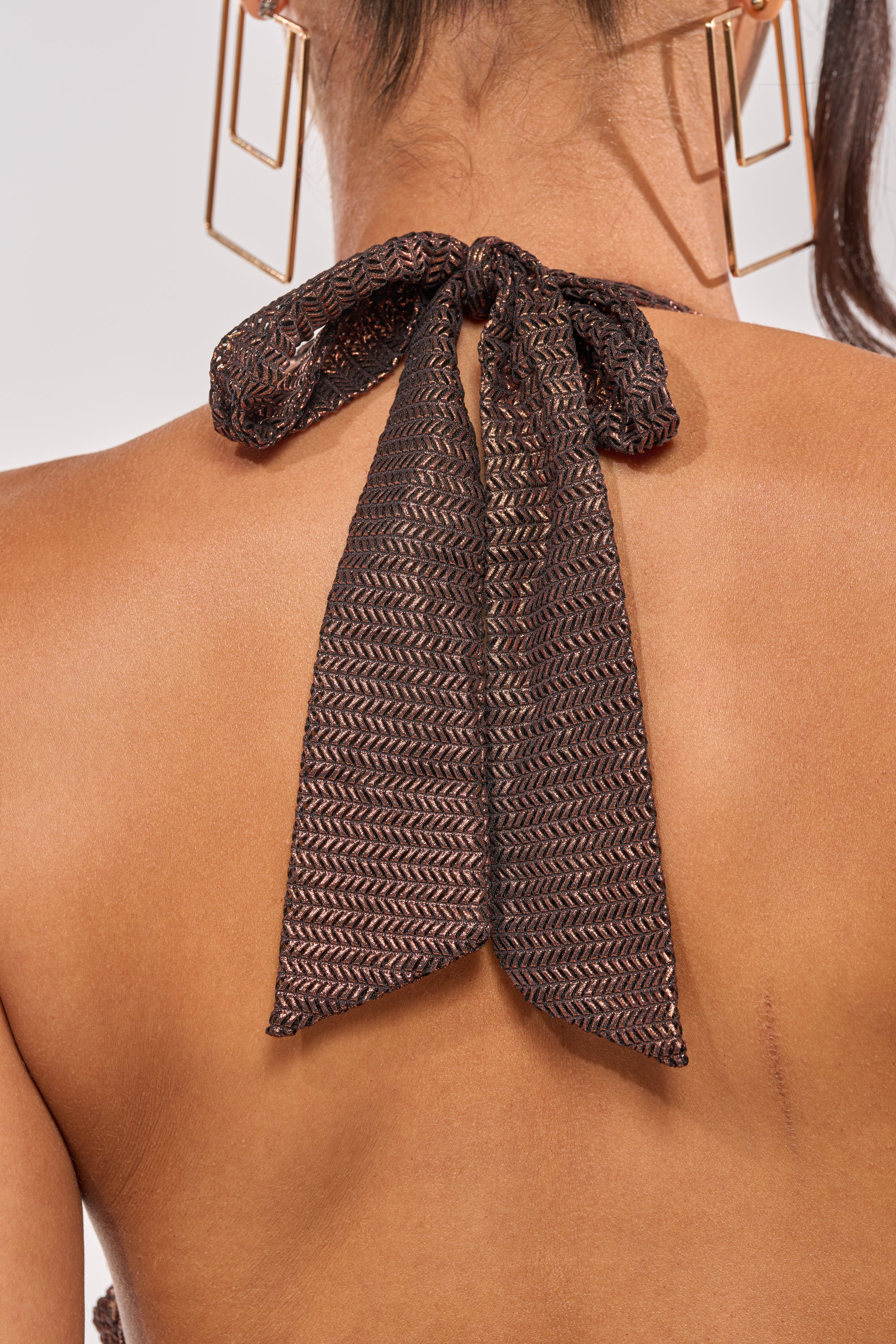 Close-up of a woman wearing the KAYLA HALTER MINI DRESS IN BROWN, featuring a textured halter tie bow at her neck. Gold geometric earrings and tied-back hair highlight the elegant neckline against a plain, blurred background. 7