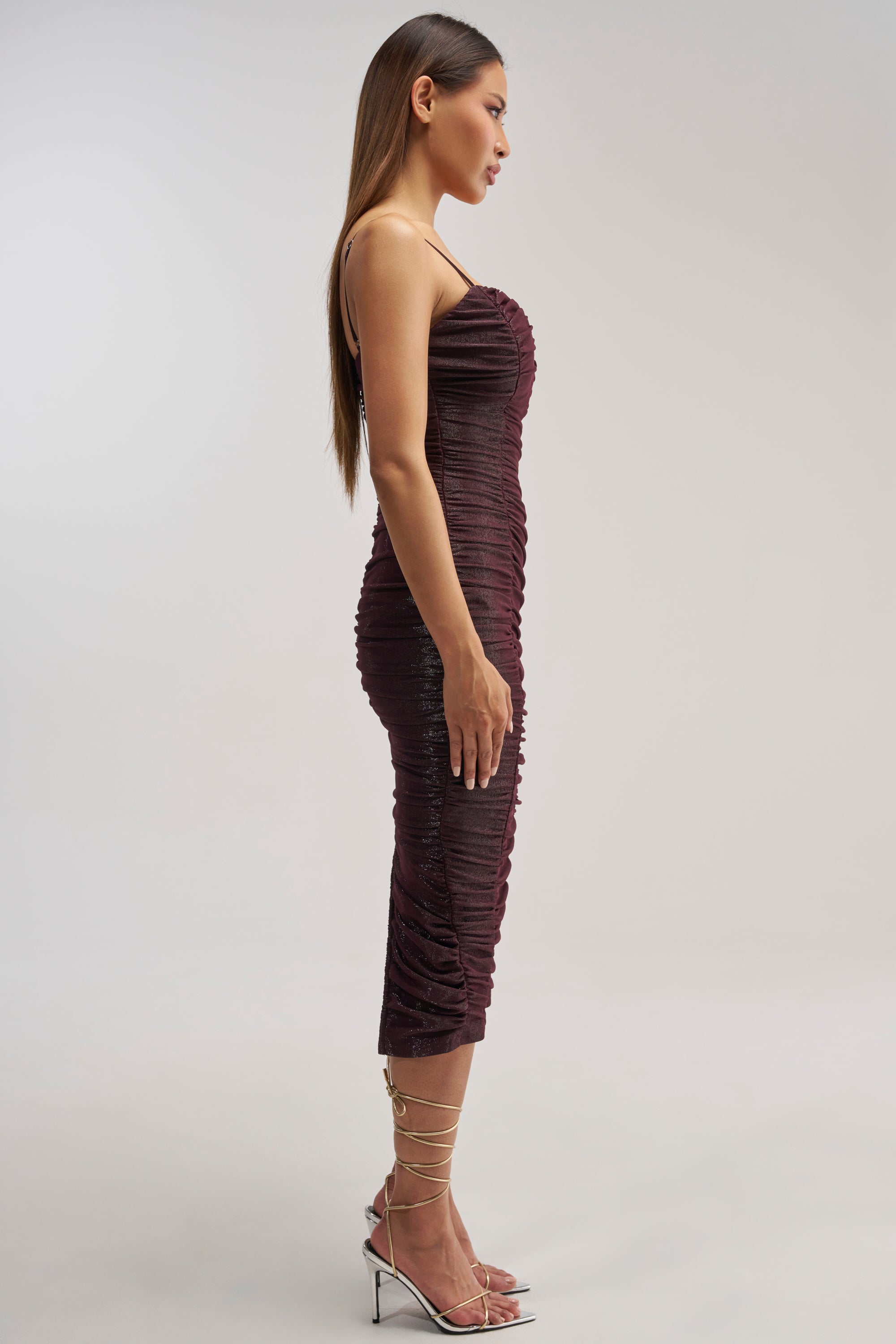 A woman stands in profile against a plain light background, wearing the FOREVER FLIRTY RUCHED MIDI DRESS—a fitted, dark burgundy bodycon style with spaghetti straps and ruching—paired with metallic strappy high-heeled sandals.