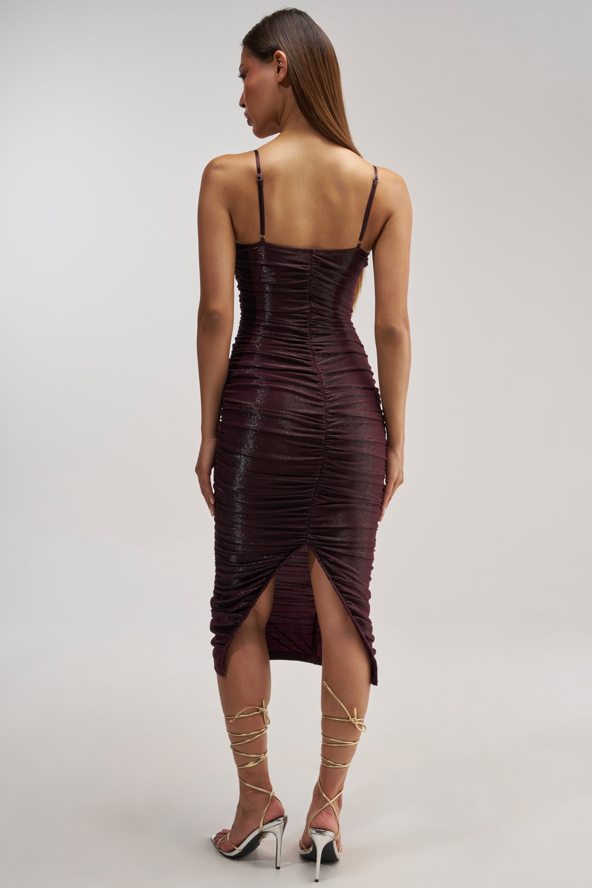 A woman stands facing away, wearing the FOREVER FLIRTY RUCHED MIDI DRESS—a fitted, sleeveless purple dress featuring elegant ruching and a back slit. She has long straight hair and completes her look with strappy gold high-heeled sandals against a plain, light-colored background.