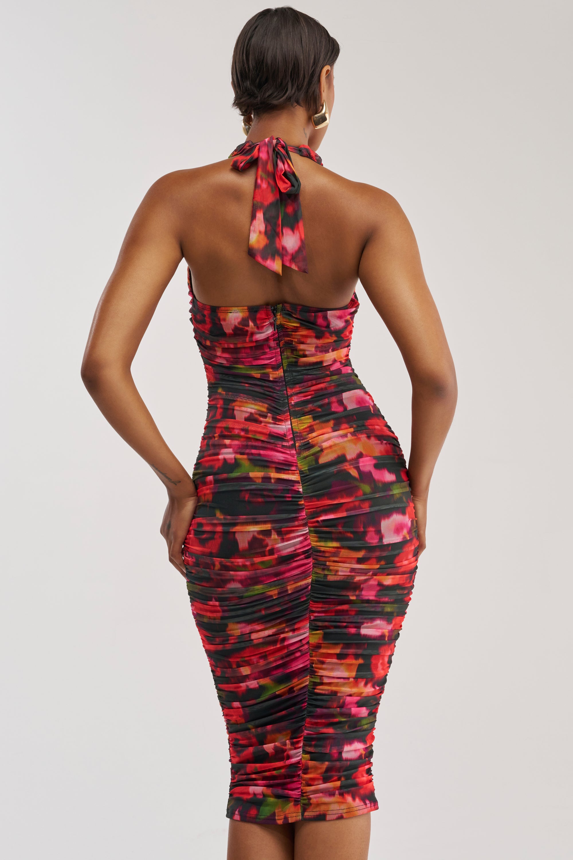 Wearing the SPELL ON YOU ABSTRACT MIDI DRESS IN PINK MULTI, a person stands facing away, showcasing the colorful fitted halter dress with its vibrant abstract print in red, pink, and orange hues. The ruched knee-length midi perfectly blends elegance and fun—ideal for a summer date night. Gold hoop earrings complement their short hair for a chic look.
