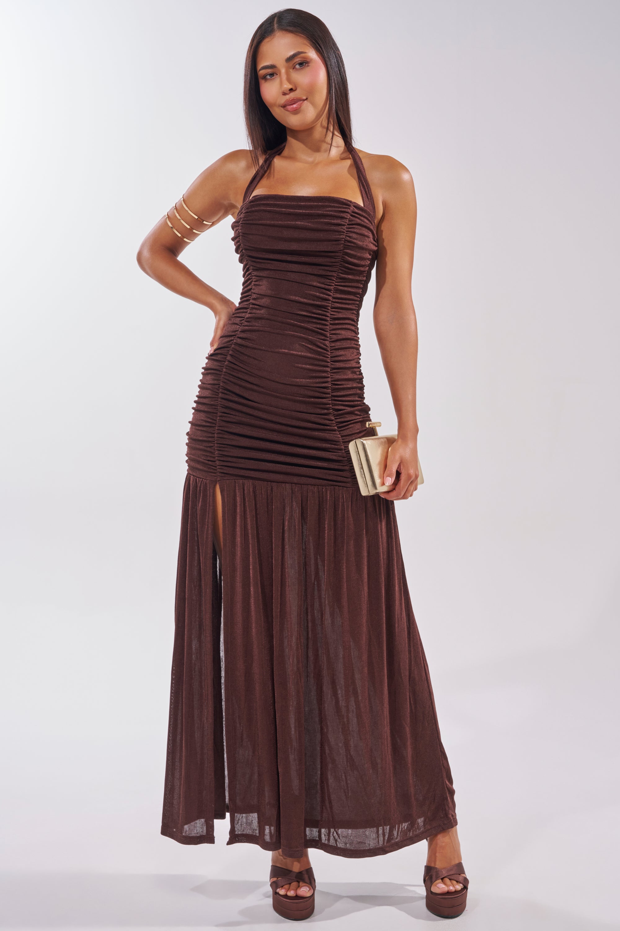 A woman stands confidently in the NOTHING SUBTLE ABOUT IT RUCHED MAXI DRESS IN BROWN with a high slit. She pairs it with a light clutch, brown platform heels, and a gold arm cuff against a plain, light background. 1