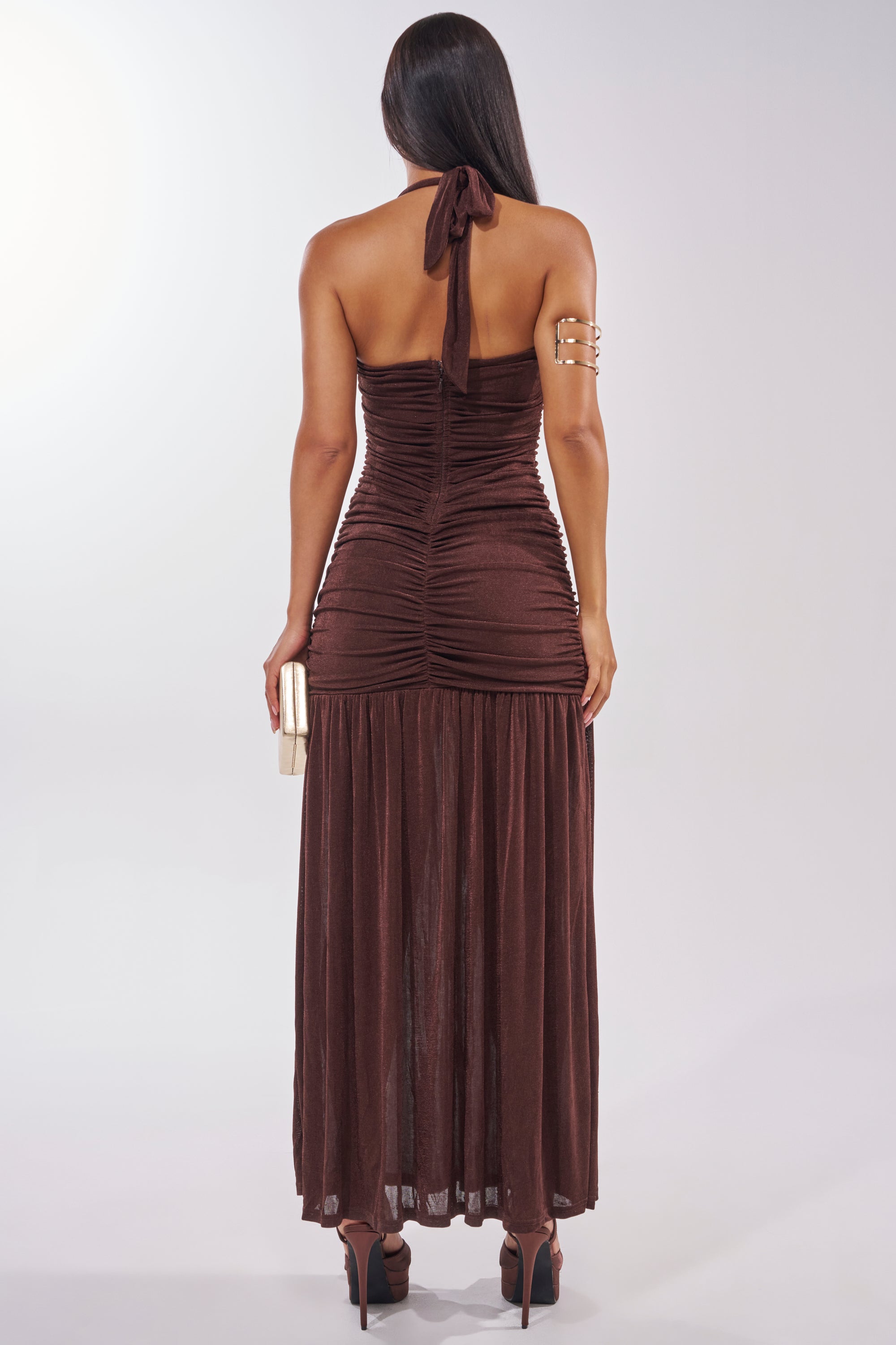 A woman with long hair faces away, wearing the NOTHING SUBTLE ABOUT IT RUCHED MAXI DRESS IN BROWN, black high heels, a gold clutch, and a gold arm cuff. 6