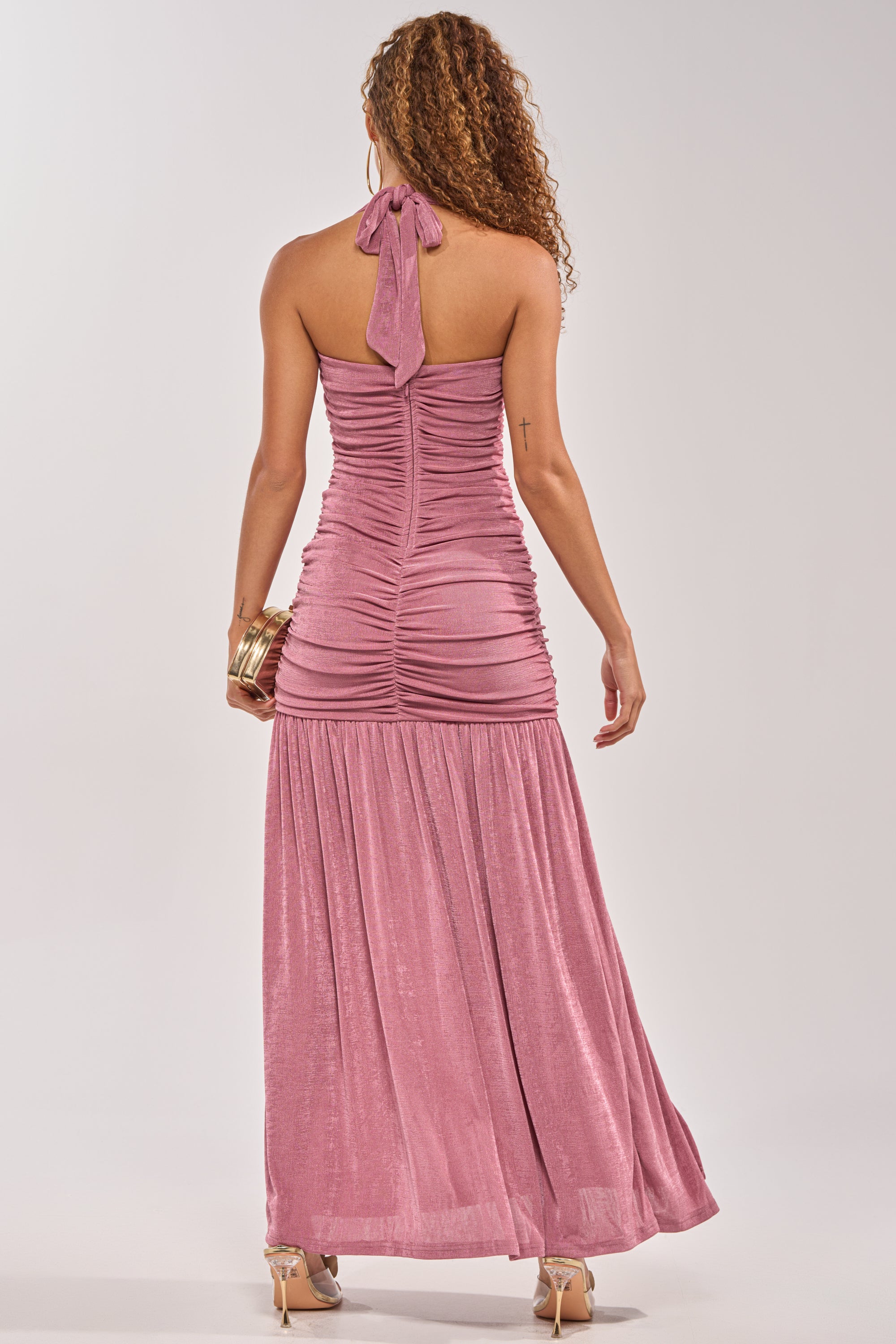 A woman with curly hair and high heels stands with her back to the camera in the NOTHING SUBTLE ABOUT IT RUCHED MAXI DRESS IN MAUVE, holding a gold clutch against a plain light background.
