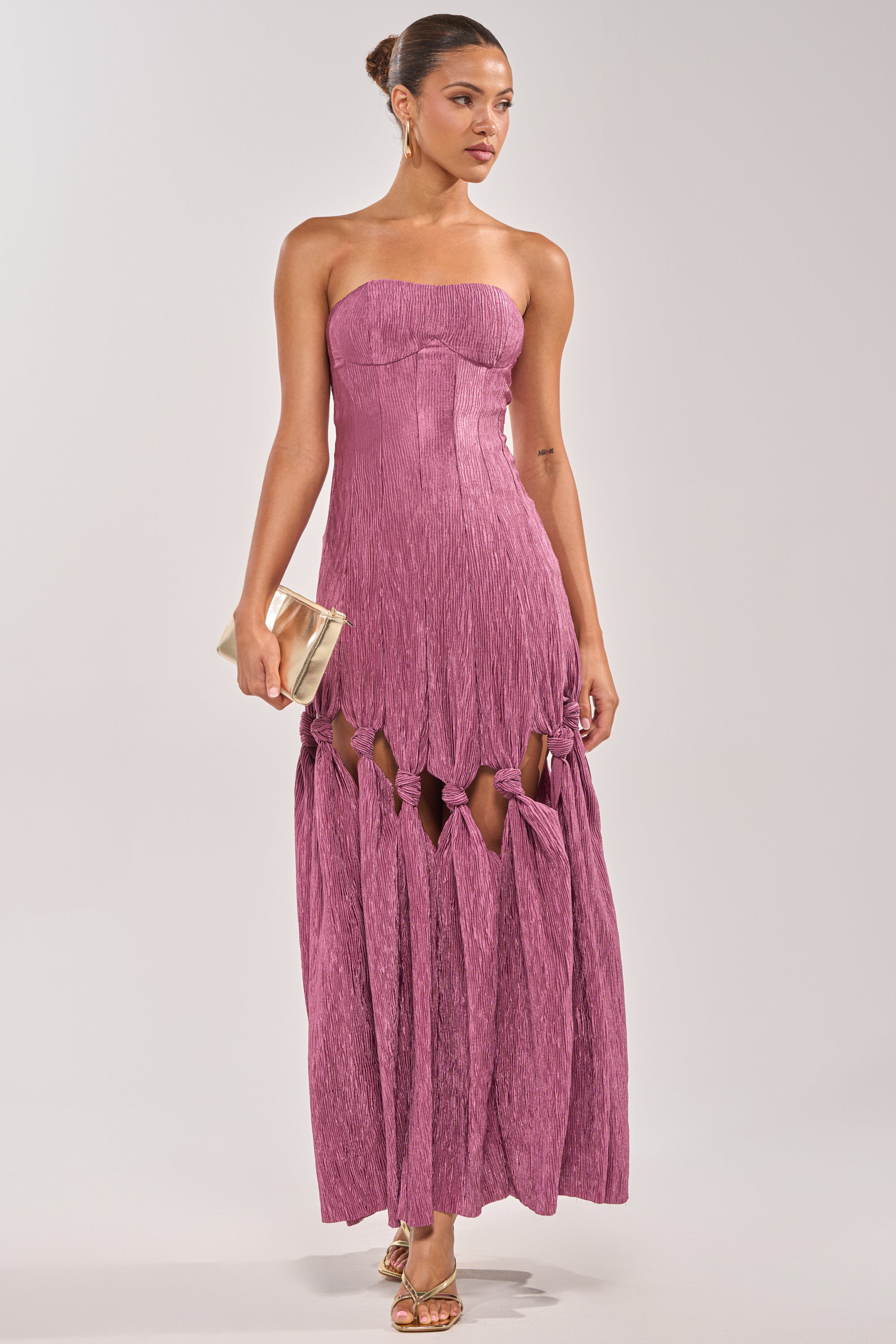 A woman in the BORN READY KNOTTED STRAPLESS MAXI DRESS—mauve, textured, with knot and cutout details—styled with gold strappy heels, a sleek bun, and a gold clutch. A chic choice for wedding guests. 0