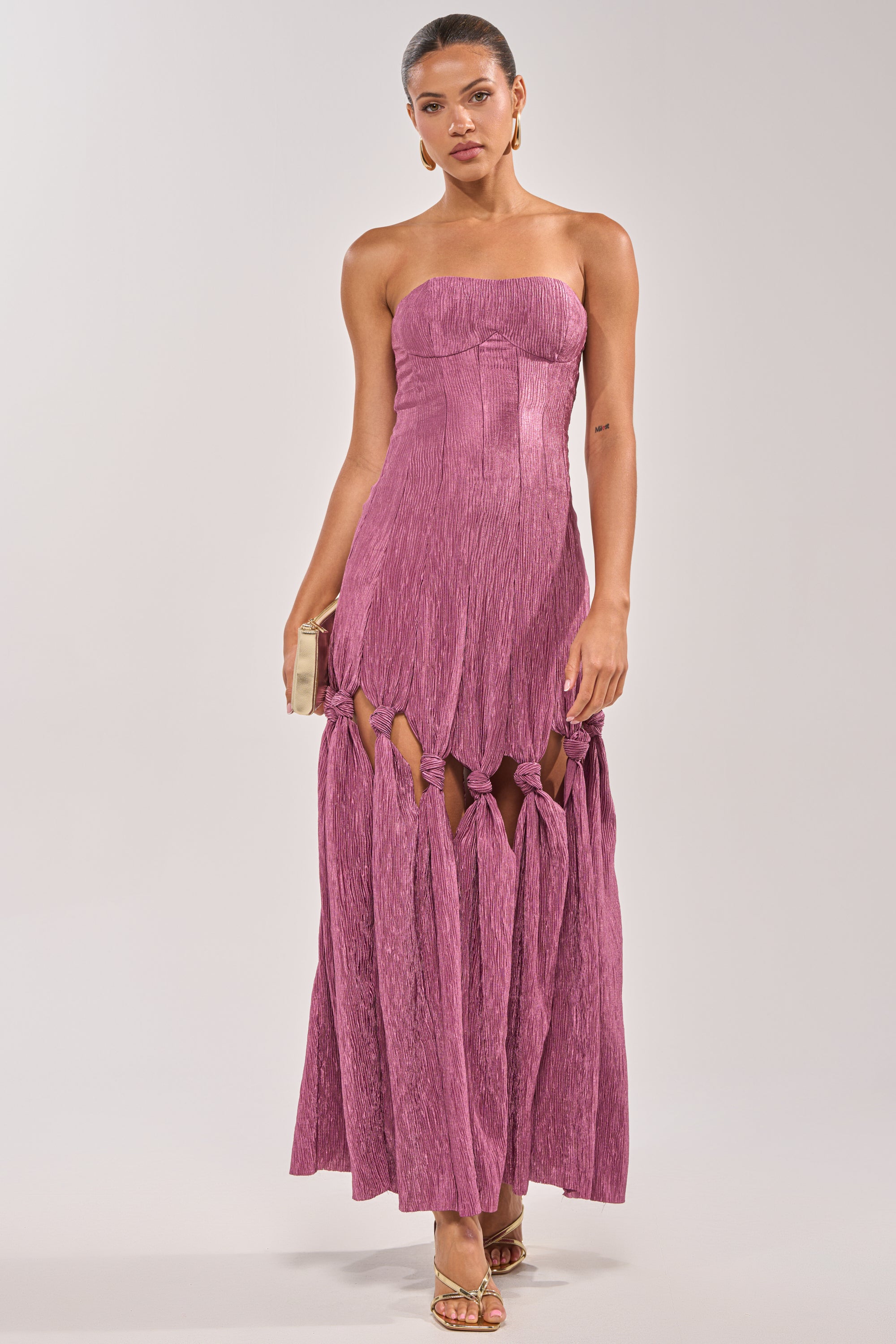 A woman in the BORN READY KNOTTED STRAPLESS MAXI DRESS—mauve, strapless, with knots above the knees—pairs it with gold hoop earrings, gold sandals, and a gold clutch for an elegant wedding guest look against a light background. 1