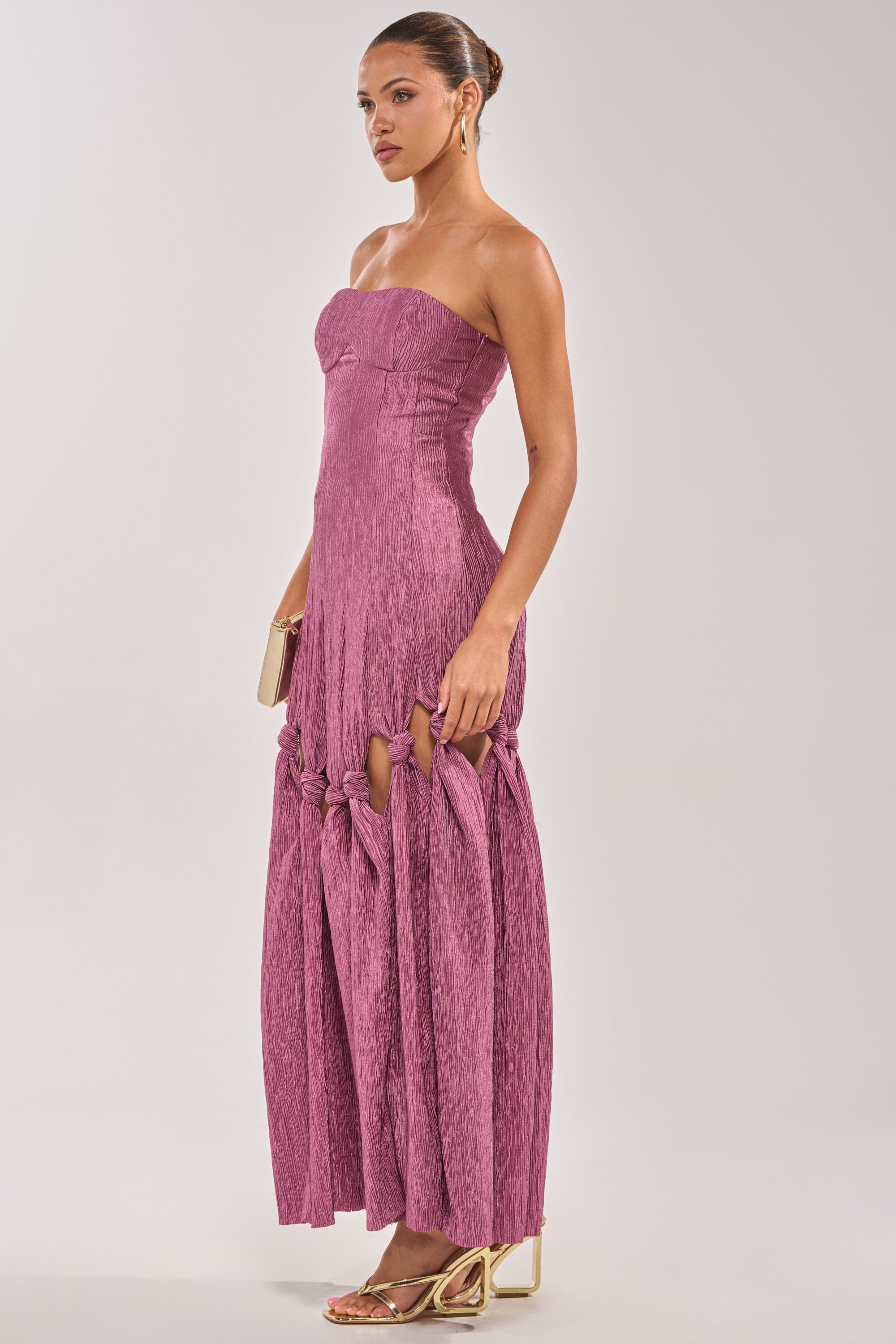 A woman in the BORN READY KNOTTED STRAPLESS MAXI DRESS—with mauve, gathered knots on the skirt—stands in profile holding a clutch. She pairs gold heels, hoop earrings, and a neat bun for an elegant wedding guest look. 2