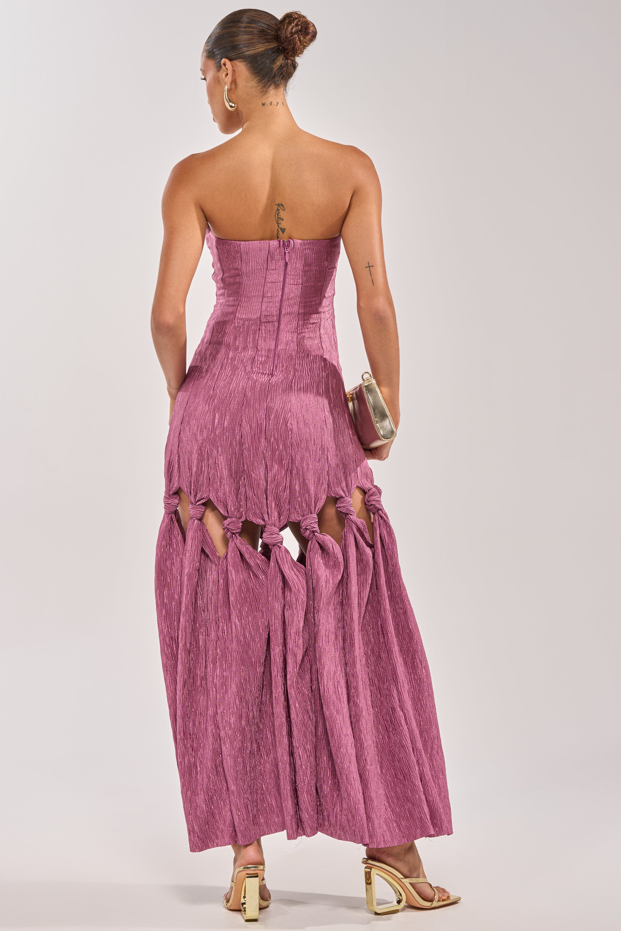 A woman faces away, wearing the BORN READY KNOTTED STRAPLESS MAXI DRESS in mauve, with knot detail cutouts on the lower skirt. She pairs it with a metallic clutch, heeled sandals, and a neat bun—ideal for wedding guests. 3