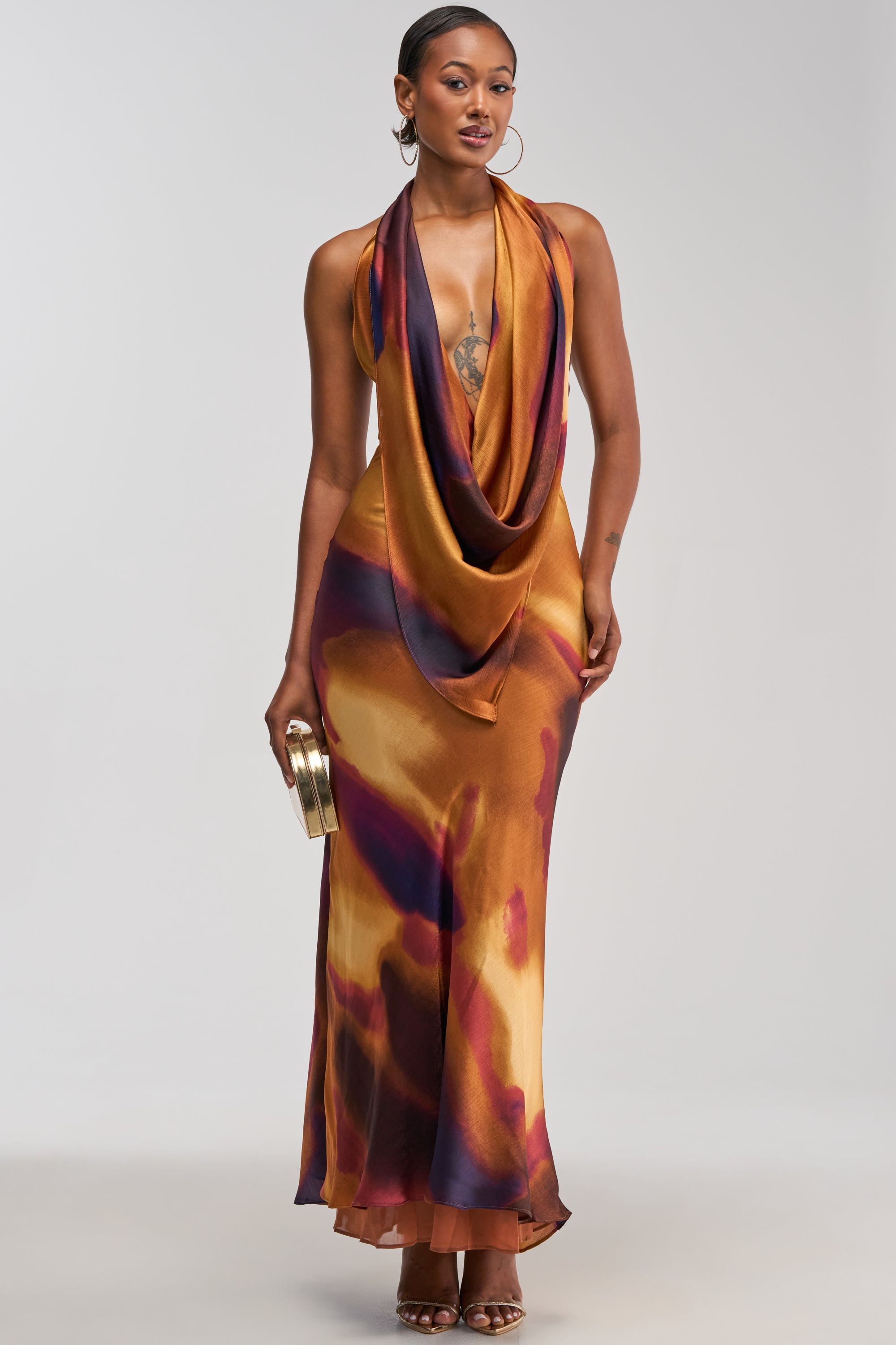 A woman wears the SUNKISSED ABSTRACT MAXI DRESS IN BROWN MULTI—a halter-neck, draped satin maxi dress in a blend of gold, orange, and purple hues—styled with a small gold clutch, hoop earrings, and heels as she stands against a plain light background, making it the perfect choice for a vacation night out.