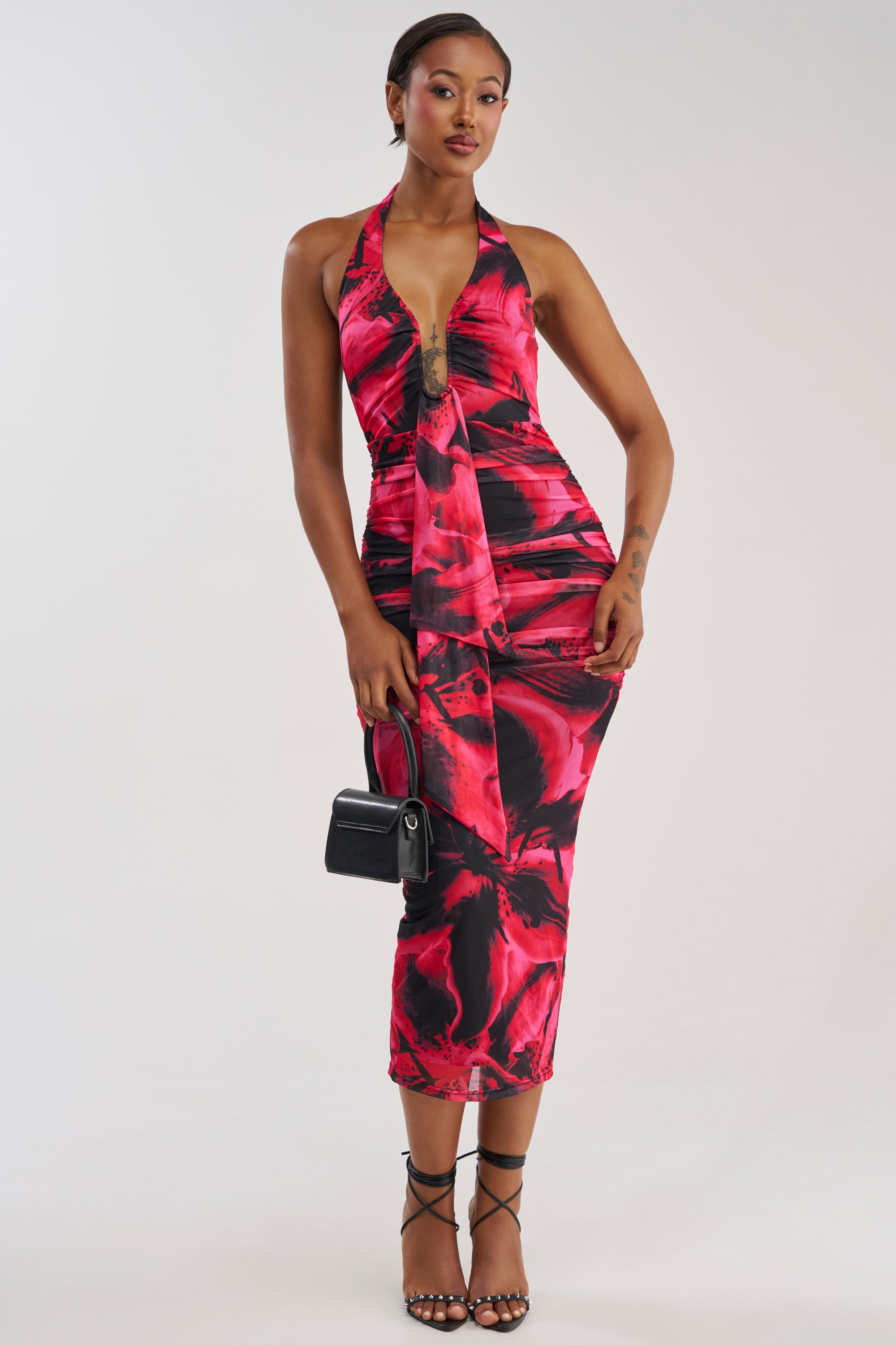 A woman wearing the MAIN STAGE ABSTRACT MAXI DRESS IN BLACK MULTI, styled with a small black handbag and black strappy heels, stands against a plain background.