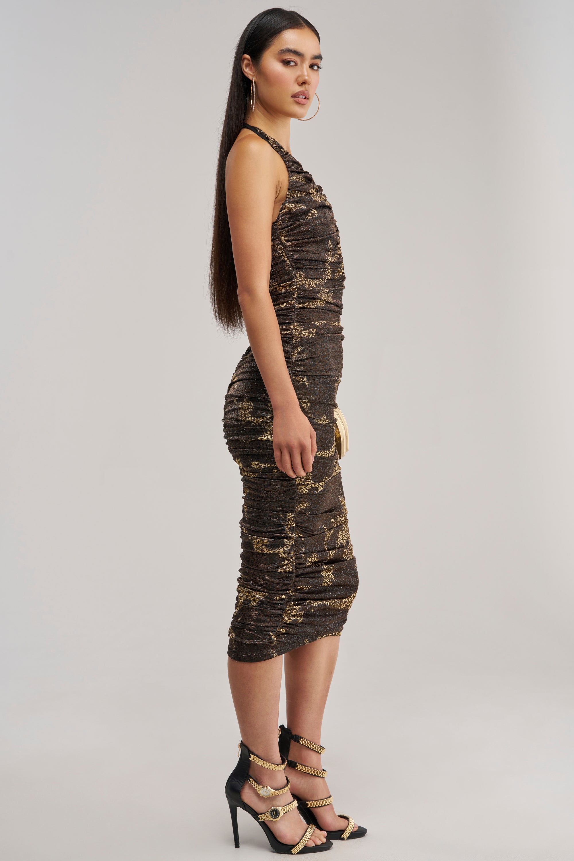 A woman stands in profile wearing the SPOTLIGHT ON ME Mesh Maxi Dress in Brown, a sleeveless, fitted dress with elegant ruched detailing and a halter neckline, paired with black high-heeled sandals and hoop earrings, against a plain light background.