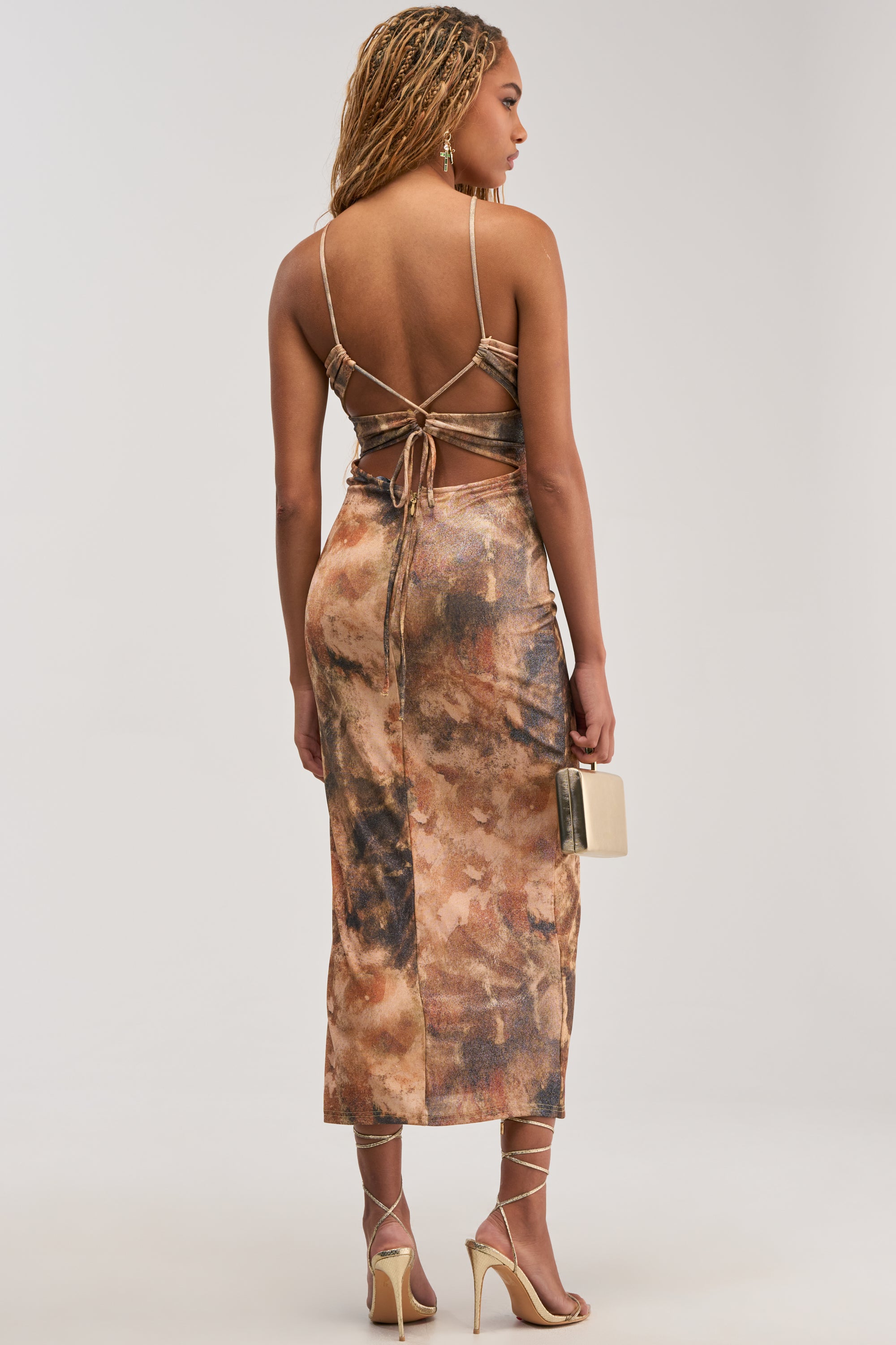 A woman with braided hair wears the CALL ME ICONIC BEADED MIDI DRESS—featuring a strappy, backless silhouette and abstract print—paired with strappy heels and a small beige clutch, standing against a plain light background.