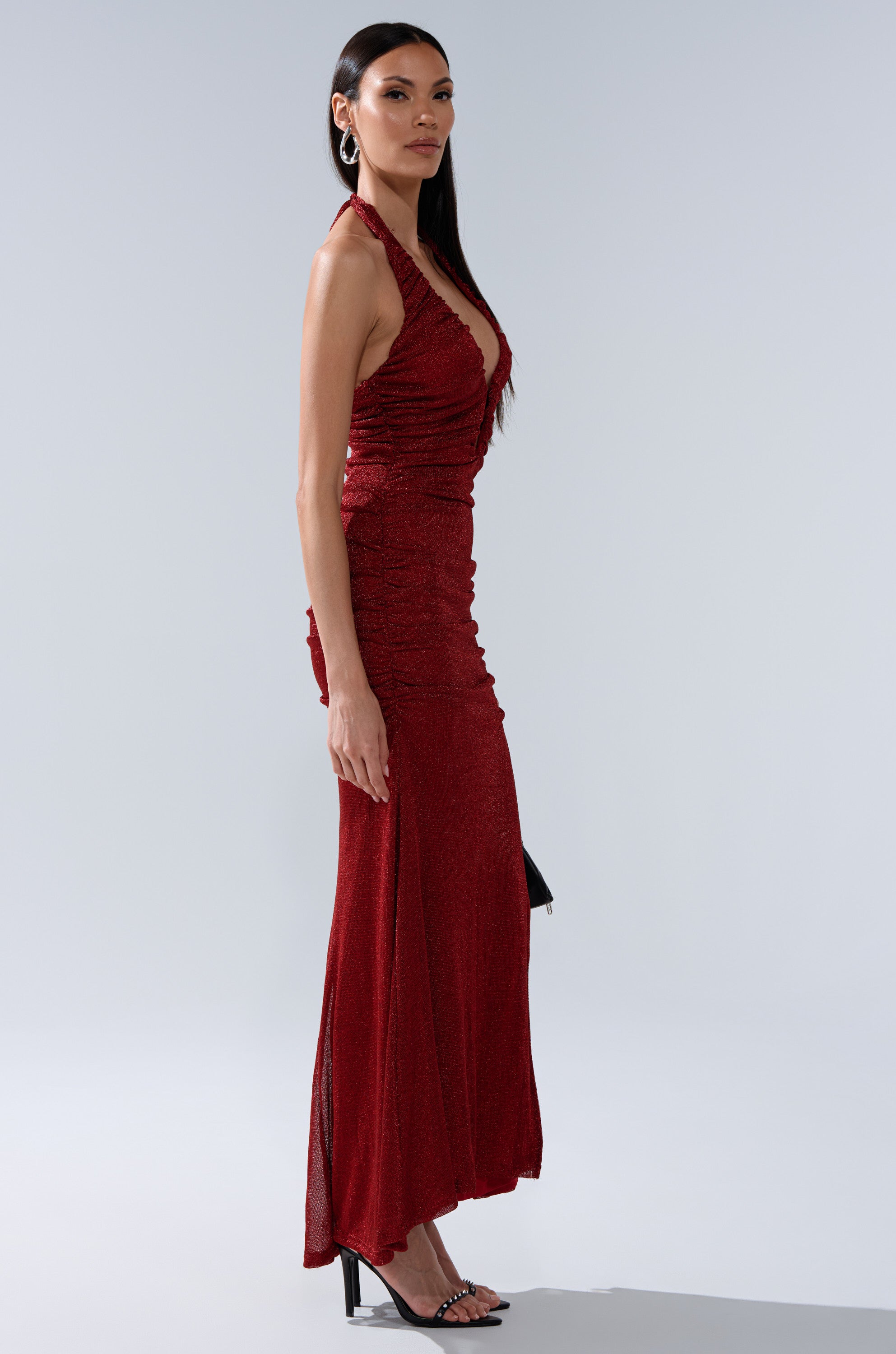 A woman in profile wears the SERPENTI PLUNGE MAXI DRESS IN METALLIC BURGUNDY—sleeveless with a deep V-neckline—paired with black heels, set against a light gray background. The shimmery knit fabric elevates her elegant look.