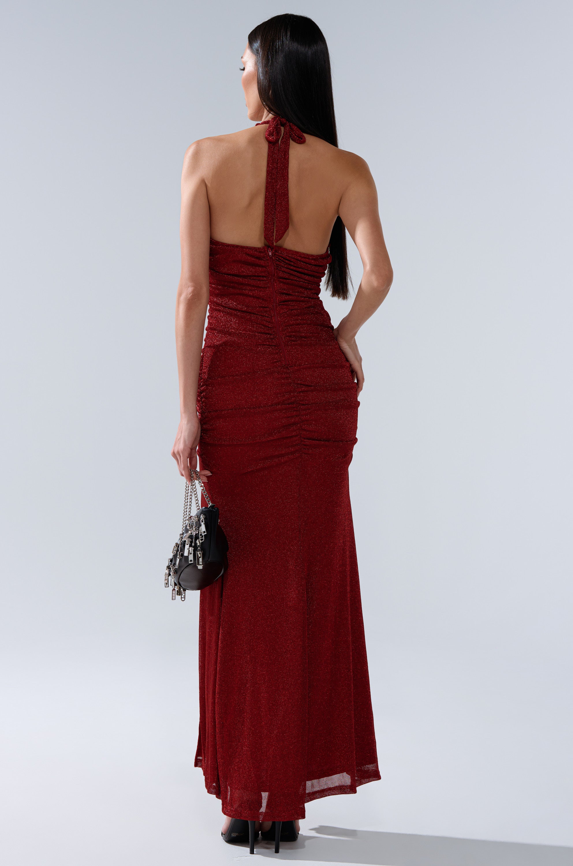 A woman with long dark hair wears the SERPENTI PLUNGE MAXI DRESS IN METALLIC BURGUNDY, showcasing its back details. She holds a small black handbag and wears black platform heels, standing against a plain background.