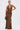 A woman in a SERPENTI PLUNGE MAXI DRESS IN METALLIC BROWN with a plunging neckline and thigh-high slit holds a small brown handbag, one hand on her hip, against a plain white background.