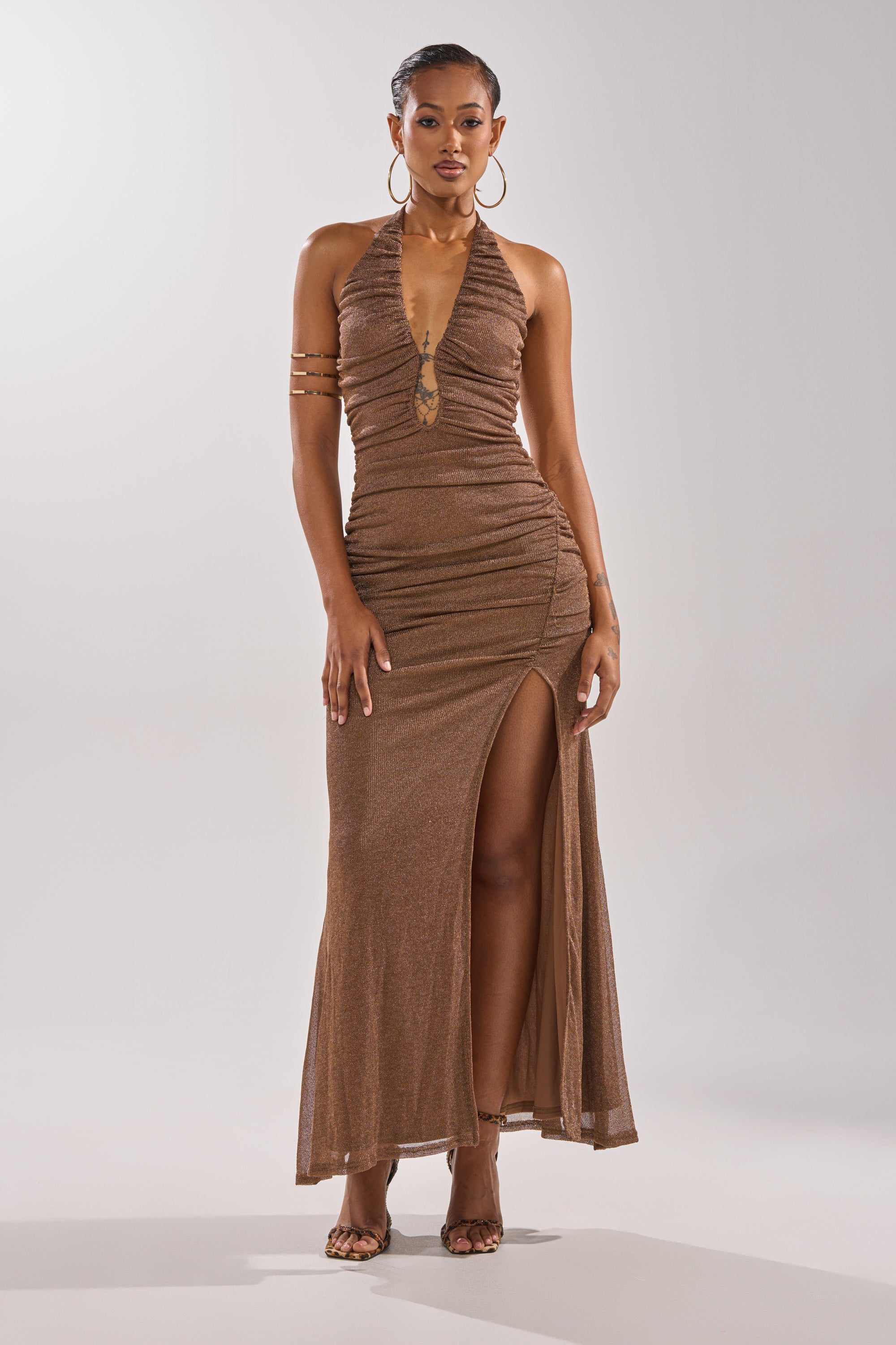 A woman poses in the SERPENTI PLUNGE MAXI DRESS IN METALLIC BROWN, featuring a sleeveless design, deep V neckline, and high thigh slit. She pairs it with strappy heels and arm jewelry against a plain light background. 0