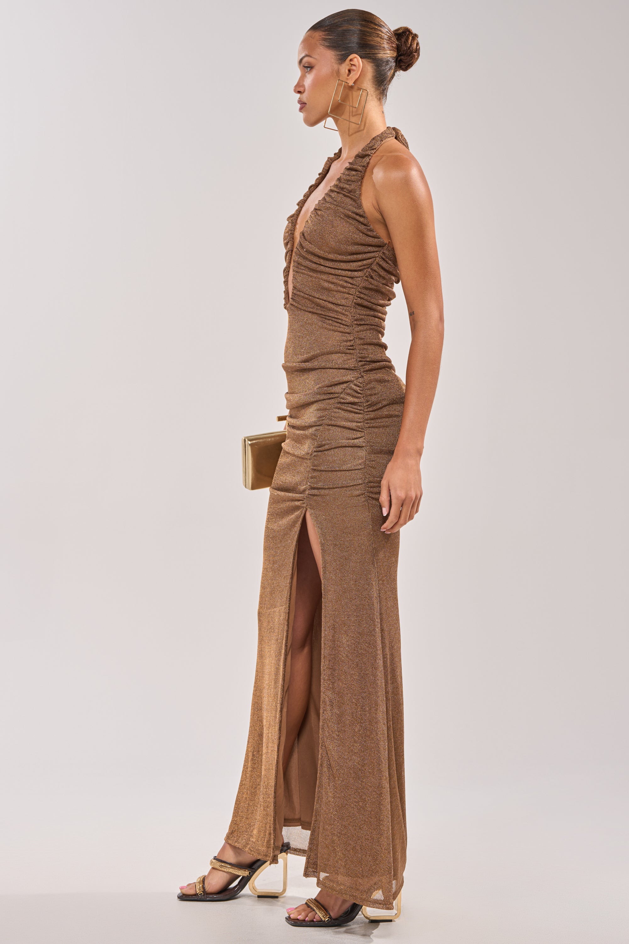 A woman in profile wears the SERPENTI PLUNGE MAXI DRESS IN METALLIC BROWN, featuring shimmery ruching and a thigh-high slit. She holds a gold clutch, with her hair in a bun, hoop earrings, heeled sandals, and a light gray background. 2