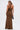 A woman with a ponytail, back to the camera, holds a small dark handbag and wears the SERPENTI PLUNGE MAXI DRESS IN METALLIC BROWN (halter neck) against a plain white background.