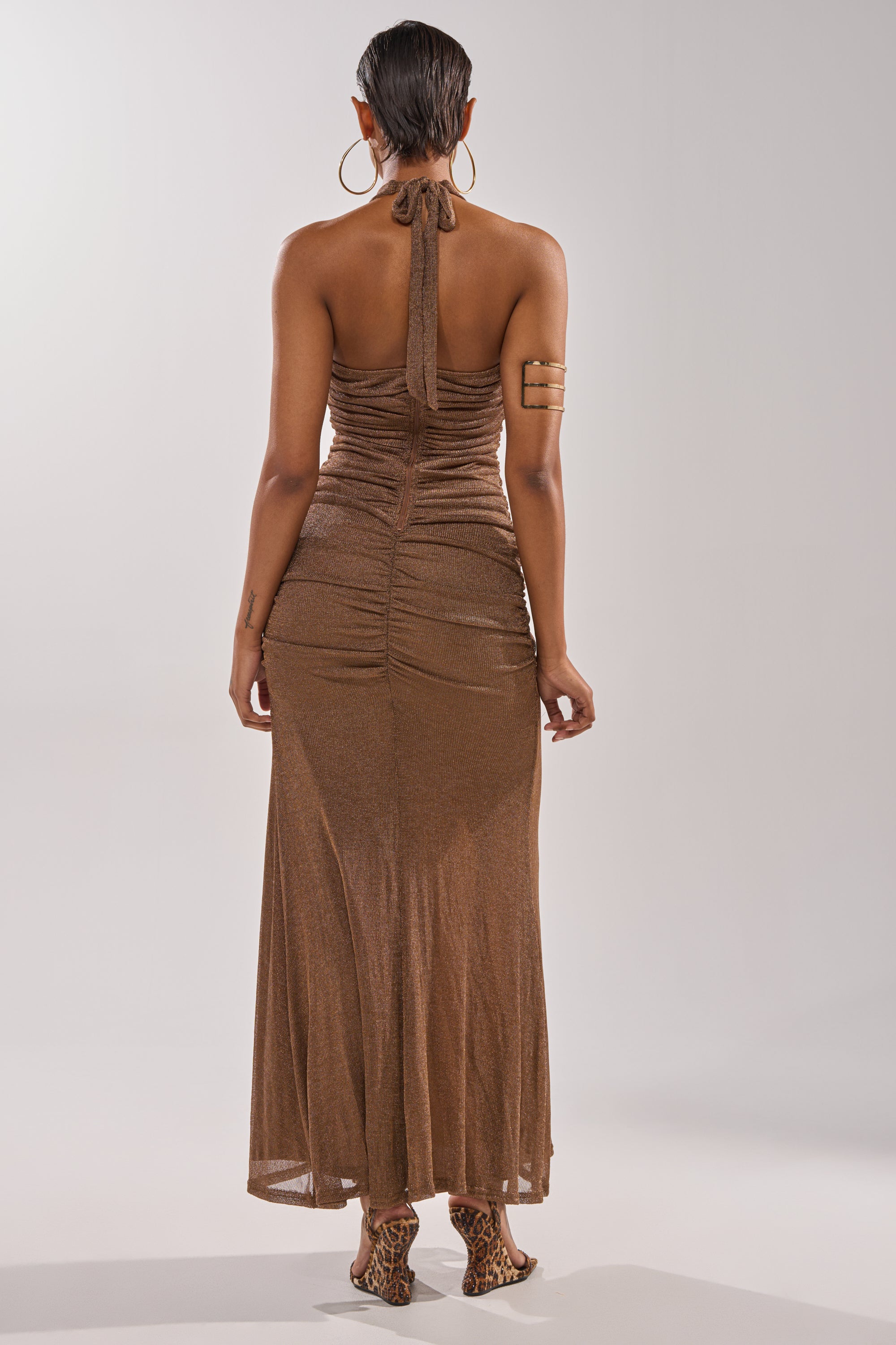 A woman faces away, wearing the SERPENTI PLUNGE MAXI DRESS IN METALLIC BROWN with a ruched back and ankle-length hem, styled with hoop earrings, an arm band, leopard-print heels, and slicked-back hair. 4