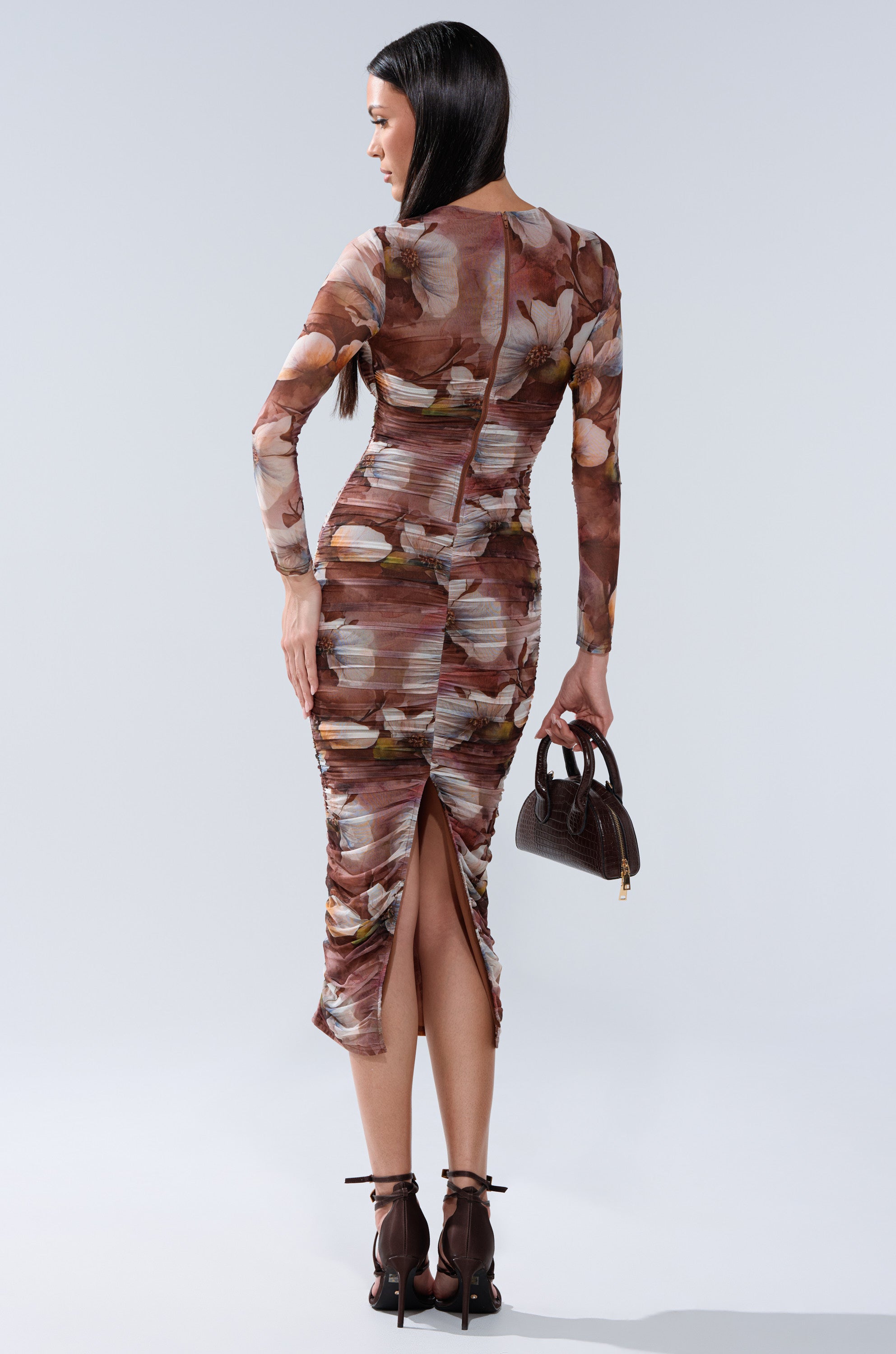 A woman with long dark hair models the FELT CUTE PRINTED MIDI DRESS, featuring a curve-hugging fit, floral print, ruched texture, and back slit. She faces away, holding a brown handbag and wearing strappy high heels. 4