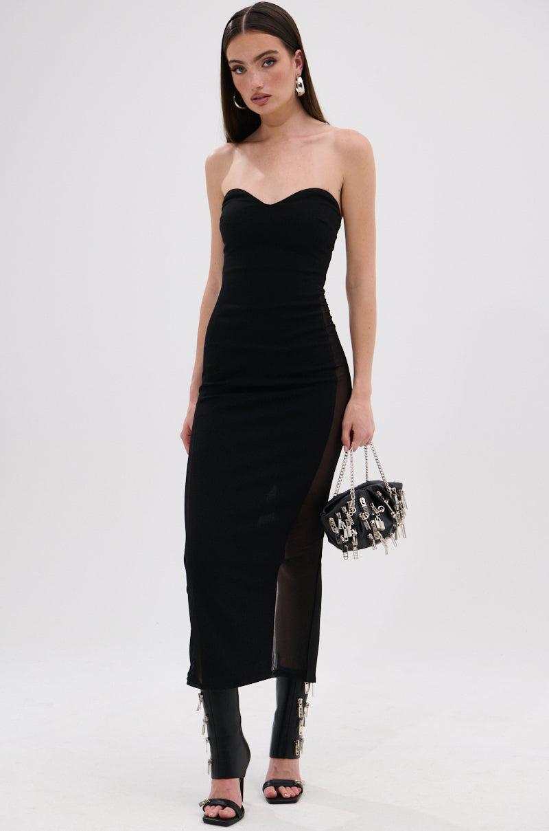 A woman in the KELLY BANDAGE MIDI DRESS, featuring a black strapless sweetheart neckline and side slit, pairs it with black open-toe heels and a small black handbag with silver embellishments against a white background. 1