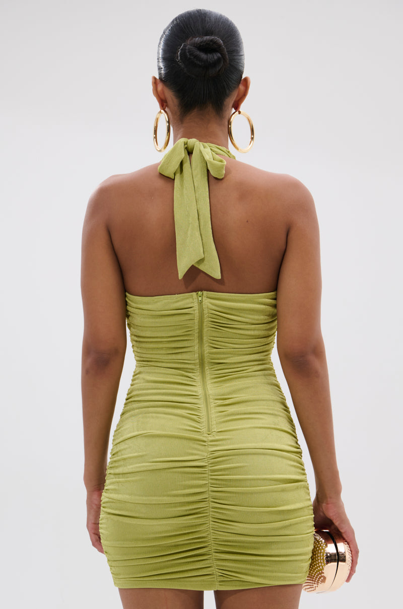 A woman is seen from behind in the green SERPENTI PLUNGE MINI DRESS with a sleek bun, holding a small round gold clutch and wearing large gold hoop earrings. 3