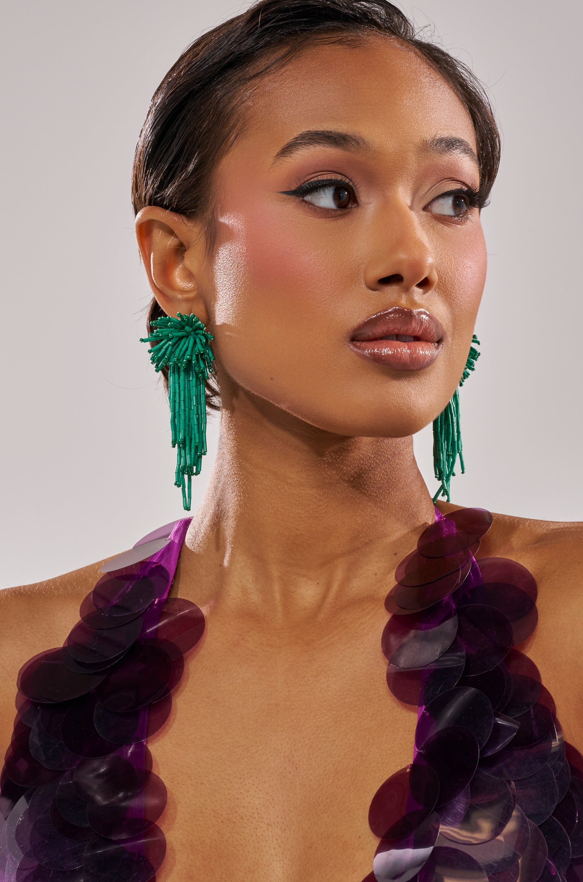 Wearing IN THE SEA EARRINGS—large green beaded statement pieces—a woman with glossy lips, winged eyeliner, and bright blush looks to the side in a top with oversized purple sequins against a neutral background. 1
