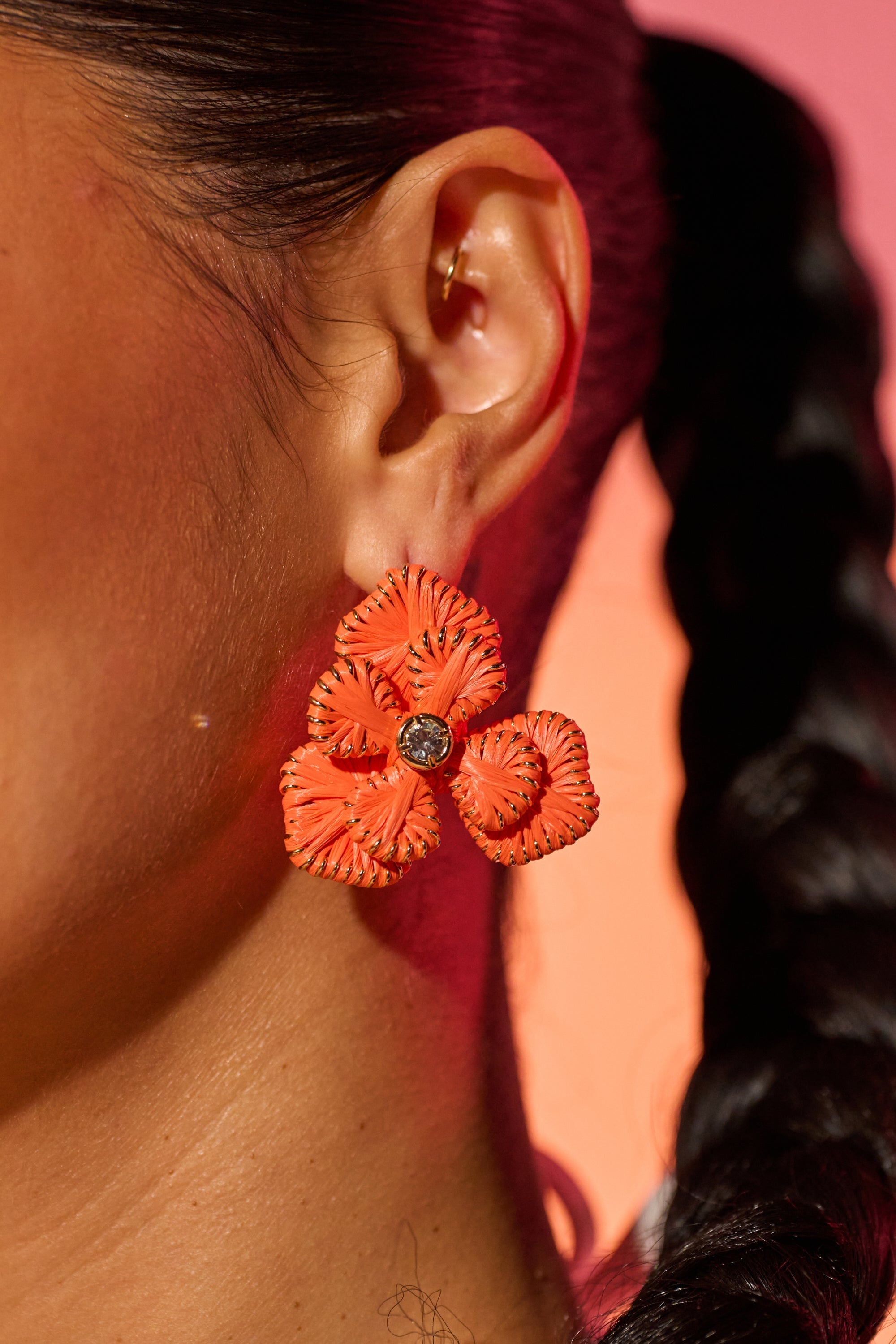 A close-up shows a person with dark braided hair wearing ON THE BEACH EARRINGS—bold statement earrings shaped like vivid orange flowers with dark centers—set against a warm pinkish-orange gradient background. 0