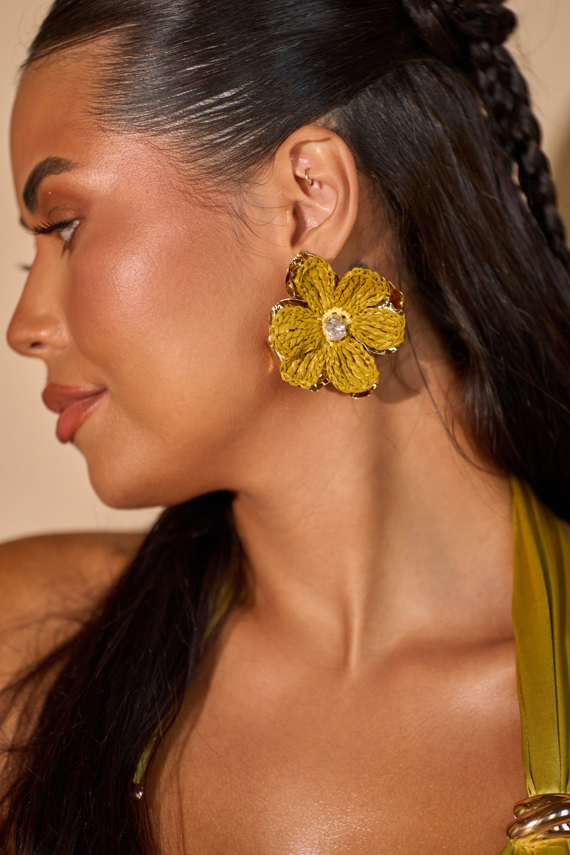 A woman with long, dark hair in braids wears the GOLDEN HOUR EARRING—bold floral statement pieces with rhinestone centers. Her skin glows naturally, and she’s dressed in a green top. 0