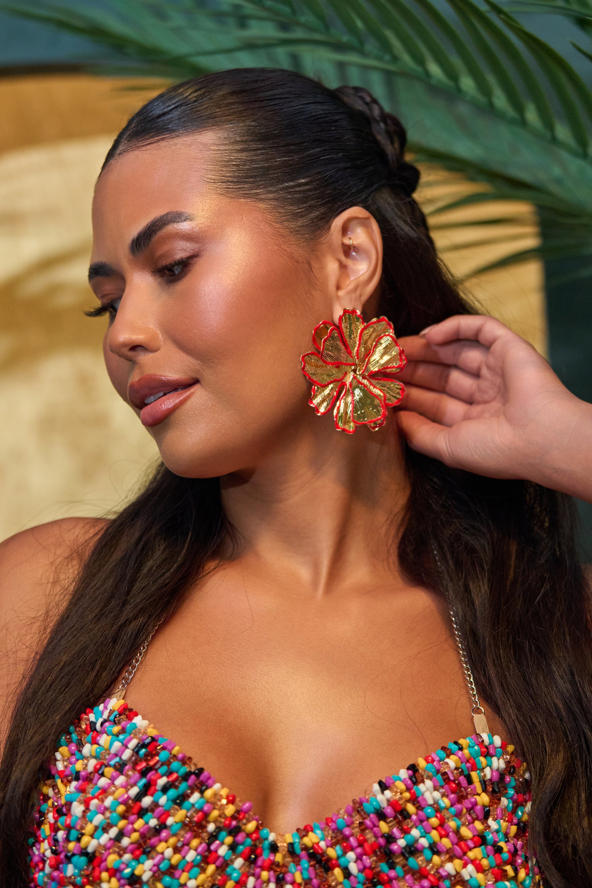 A woman with long dark hair wears the IN THE GARDEN EARRING—oversized flowers in red and metallic gold—pairing them with a colorful beaded top. She poses by a green palm leaf, capturing vibrant summer beach jewelry vibes.