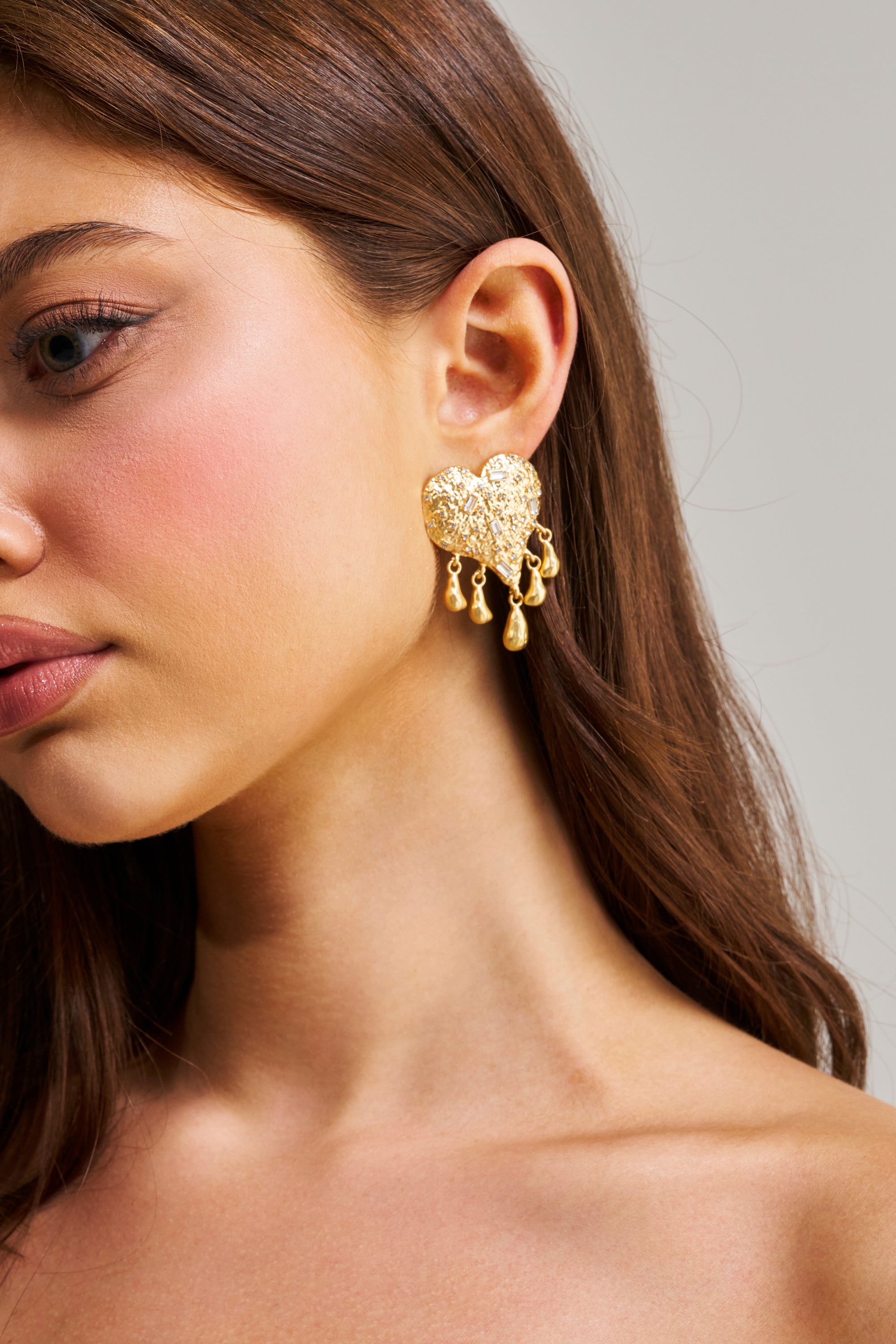 A woman with long brown hair wears the ALL MY LOVE EARRING—gold statement earrings featuring a textured heart and dangling drops. The focus is on her ear and jewelry, set against a neutral background.