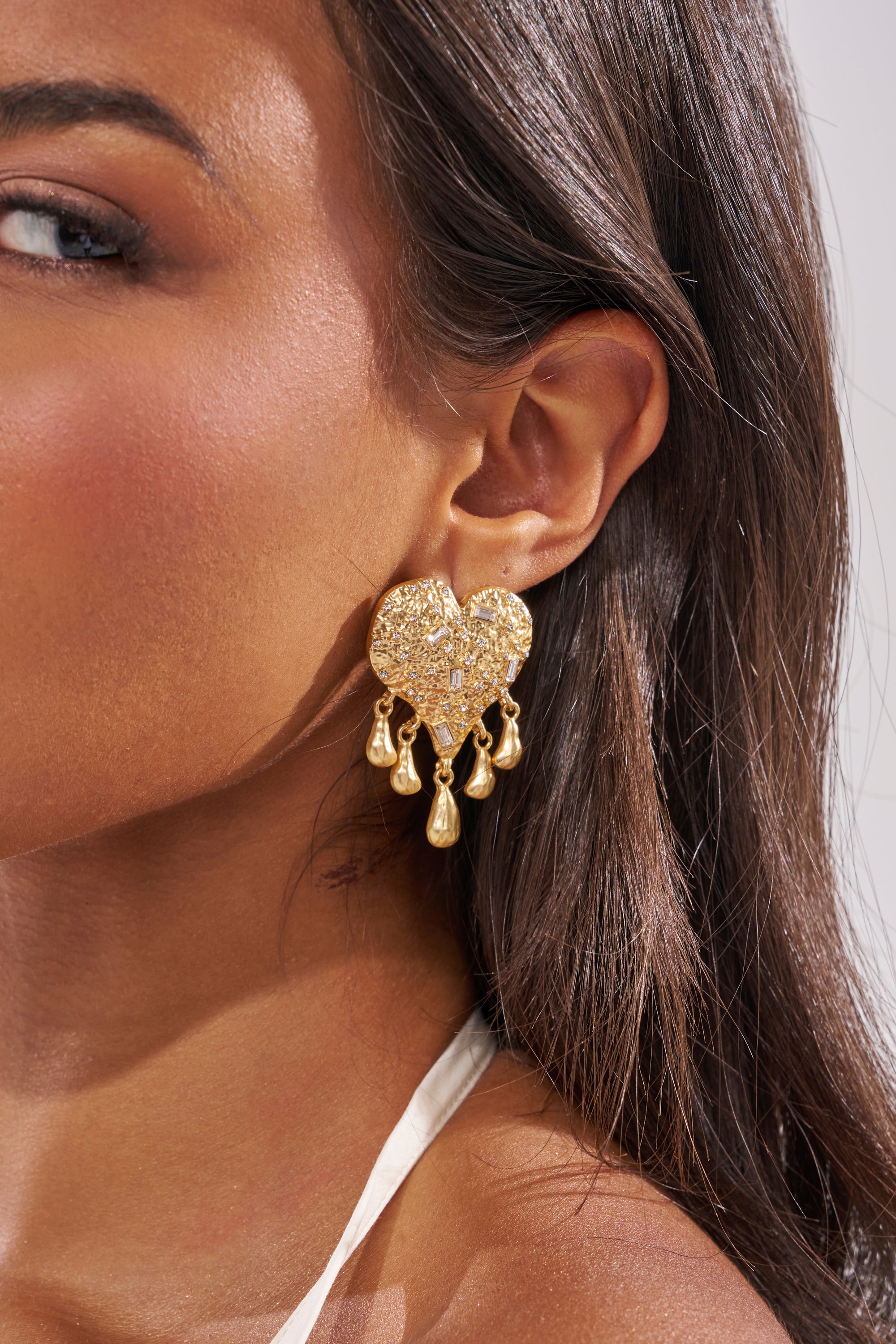 Close-up of a woman's ear wearing the ALL MY LOVE EARRING, featuring statement gold hearts with a textured surface and dangling teardrop charms. The neutral background highlights these standout earrings. 1