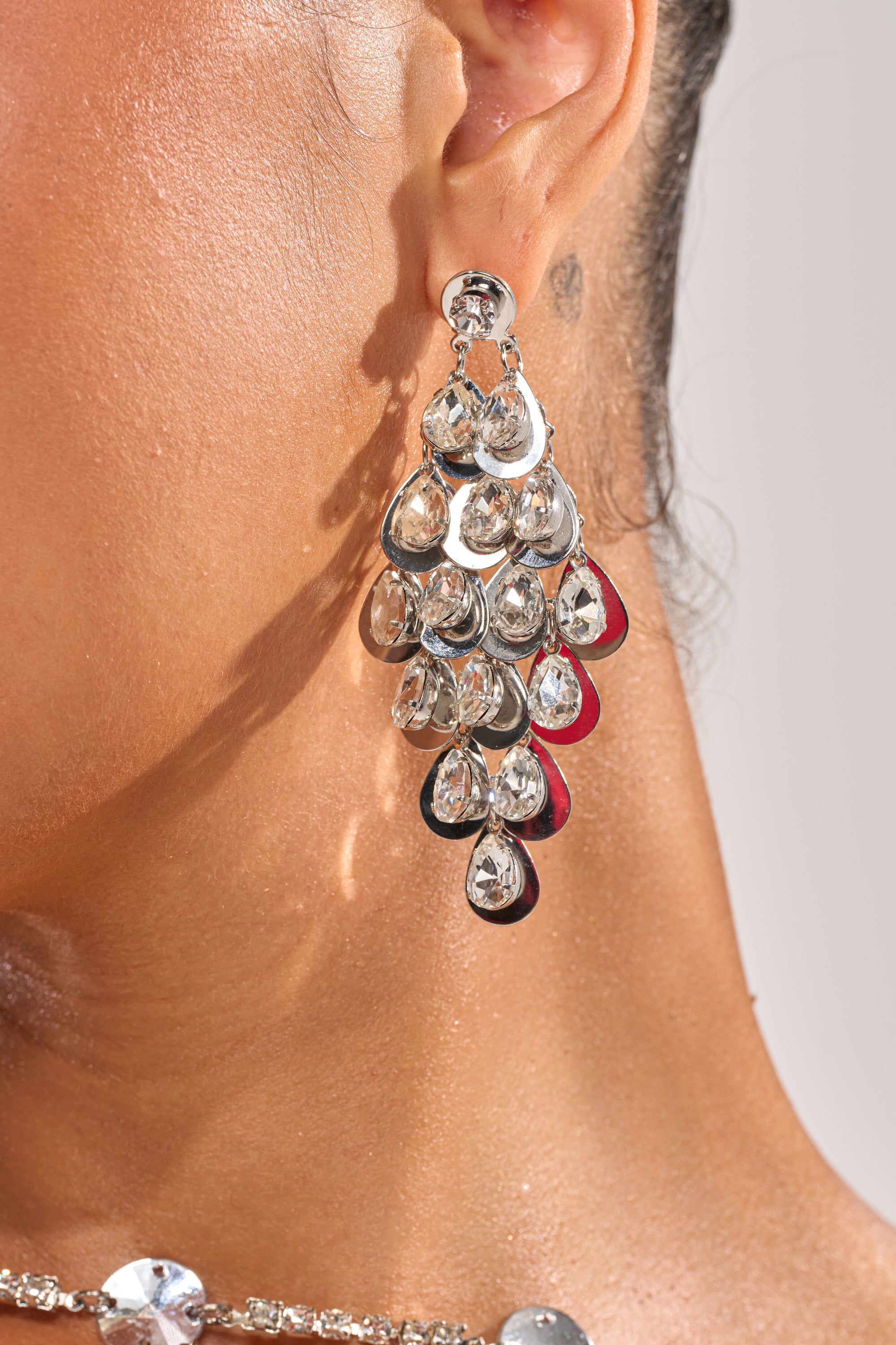 A close-up of a person’s ear wearing the DAZZLE ME EARRING, featuring rhinestone chandelier teardrop gemstones, with part of a matching necklace visible. 2