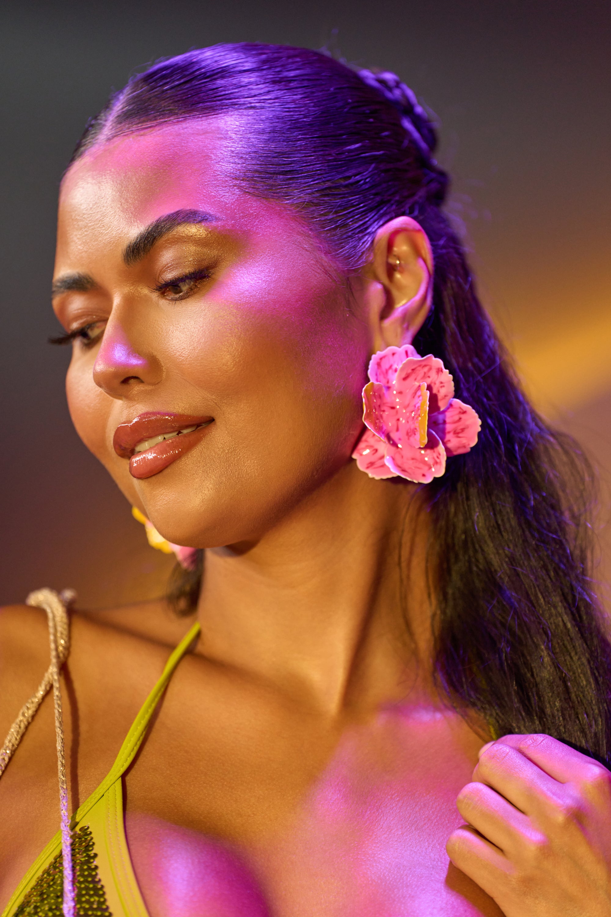 A woman with long dark hair, wearing the IM SPOILED EARRING and a green top, smiles softly while vibrant purple and yellow light reflects off her festival jewelry.