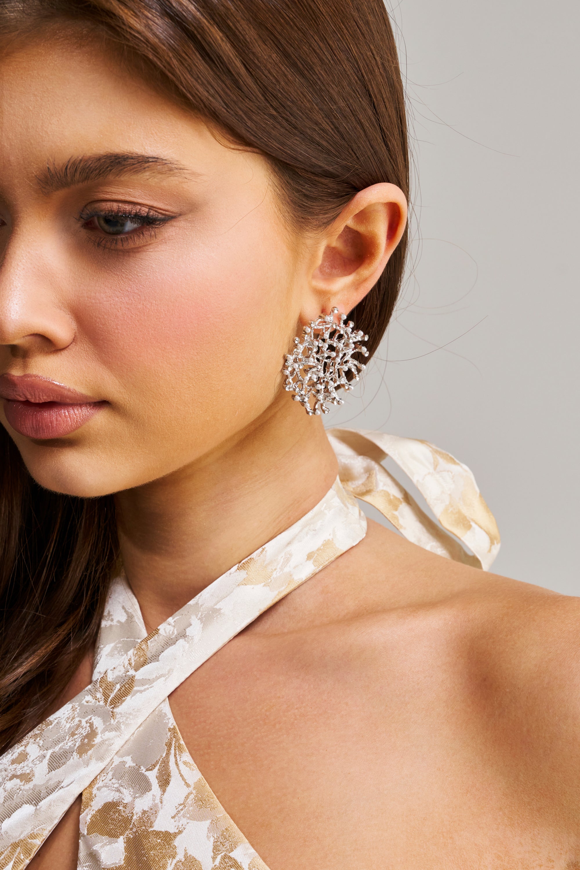 A woman with long brown hair models the TO THE REEF EARRING—large, intricate silver earrings—paired with a white patterned halter dress. The image is a shoulder-up shot, highlighting her earring and soft makeup.