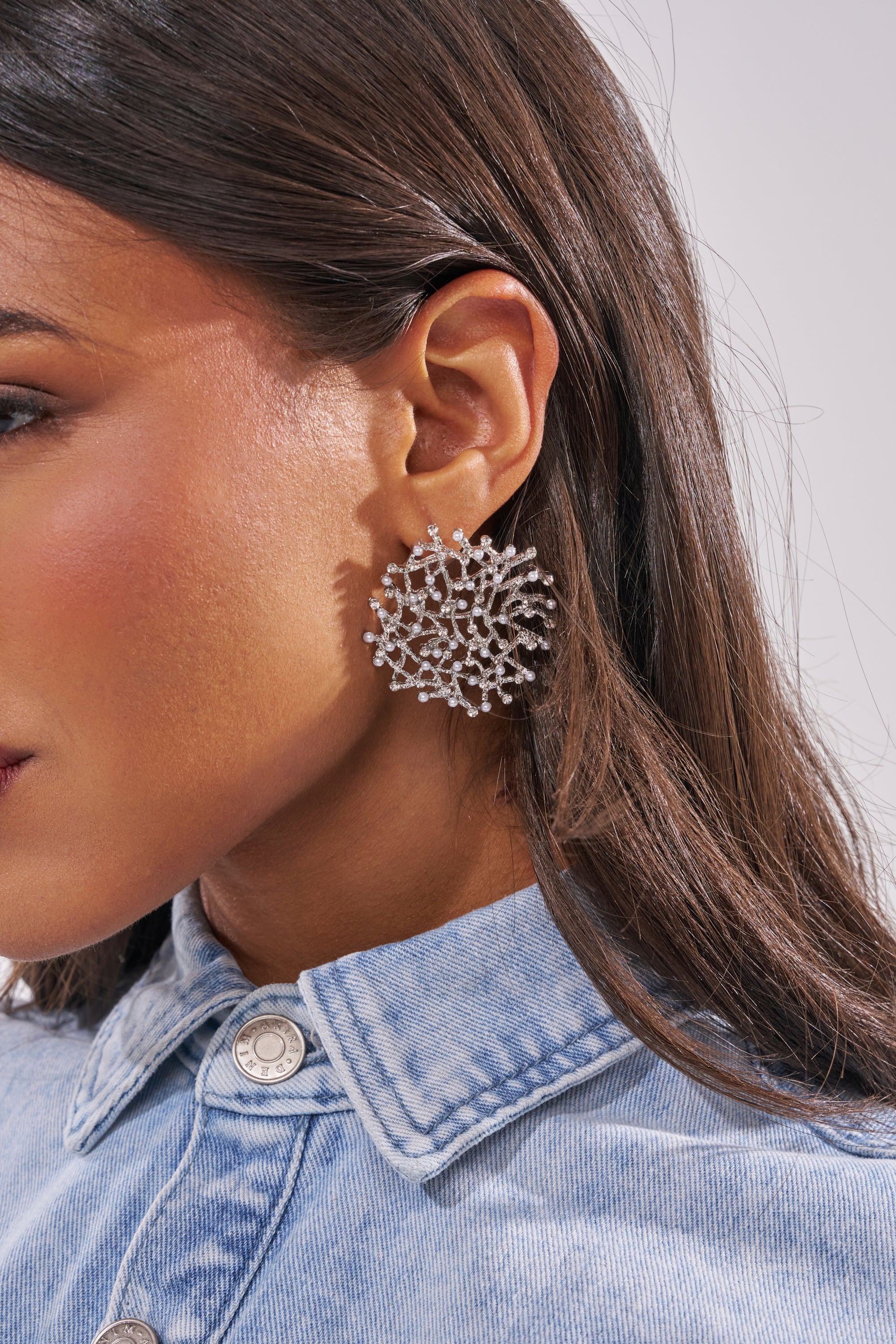 A woman with straight brown hair wears a light denim shirt and TO THE REEF EARRING, a large, intricate silver coral reef-inspired piece. The photo highlights her ear and this statement earring. 1